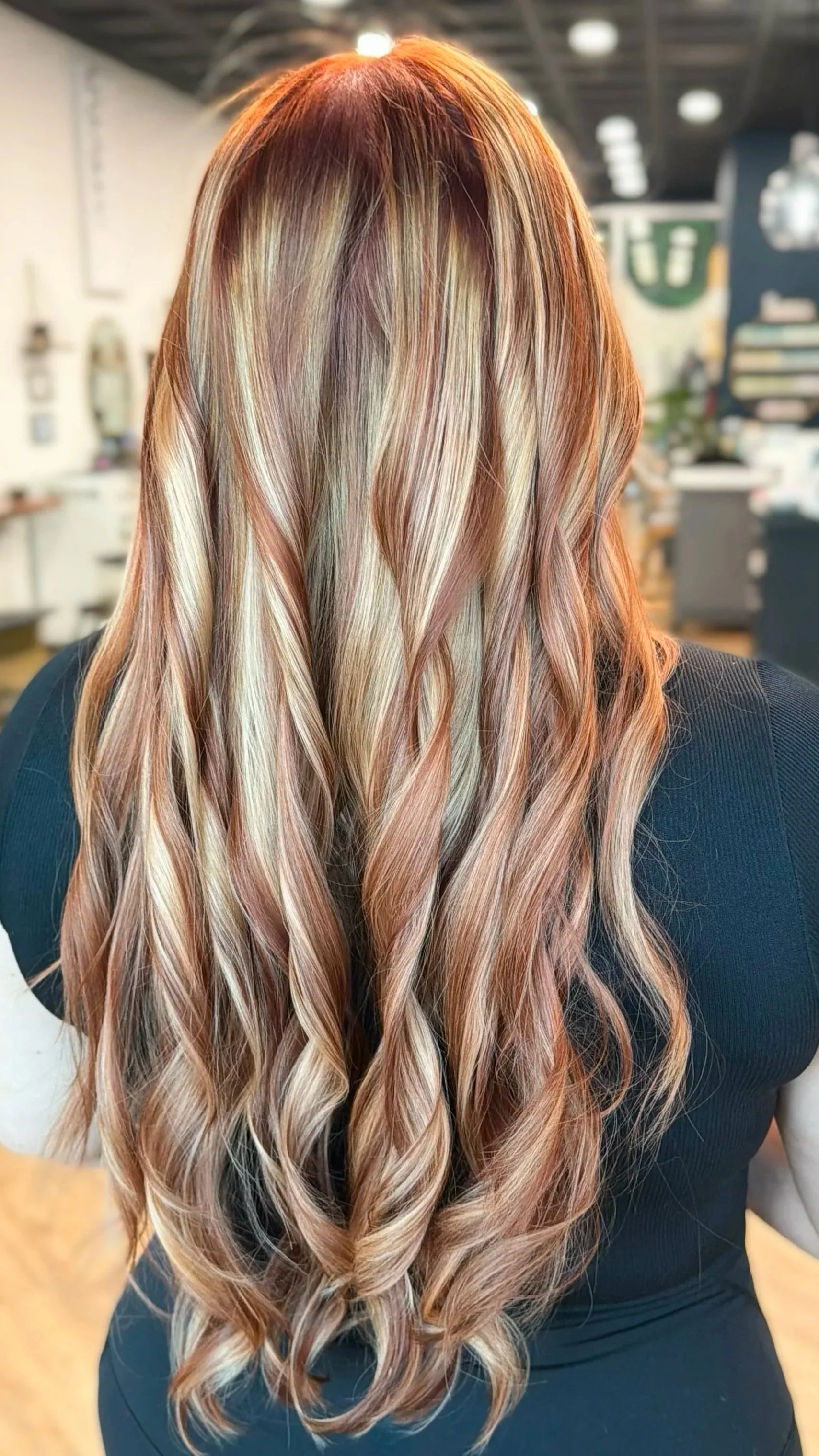 Back view of a woman with long, wavy hair styled in loose curls. Her hair features a blend of blonde and light brown shades with copper red and honey blonde chunky highlights. 