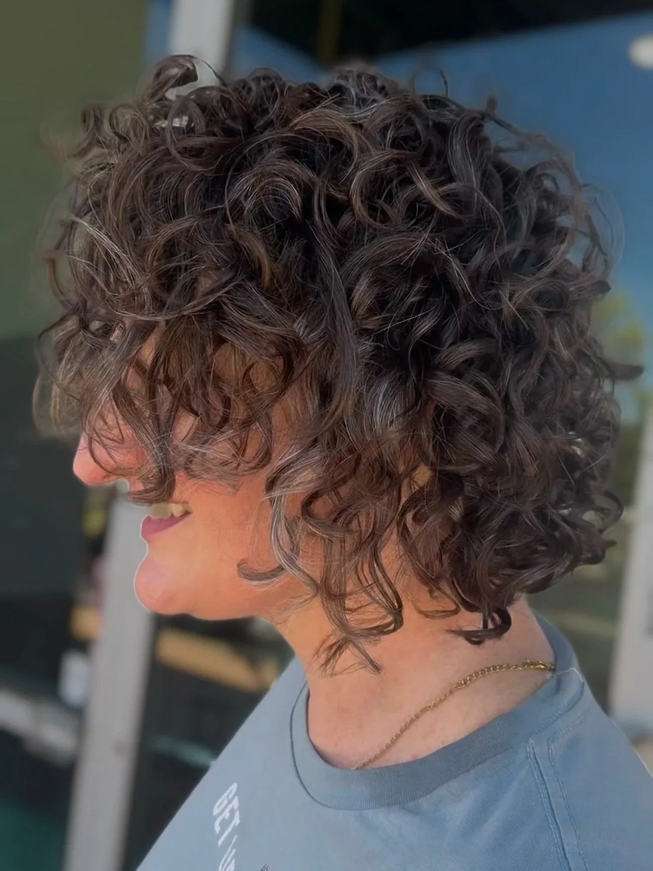 Side view of a woman with short, curly brown hair, smiling, wearing a gray shirt and a thin gold necklace.