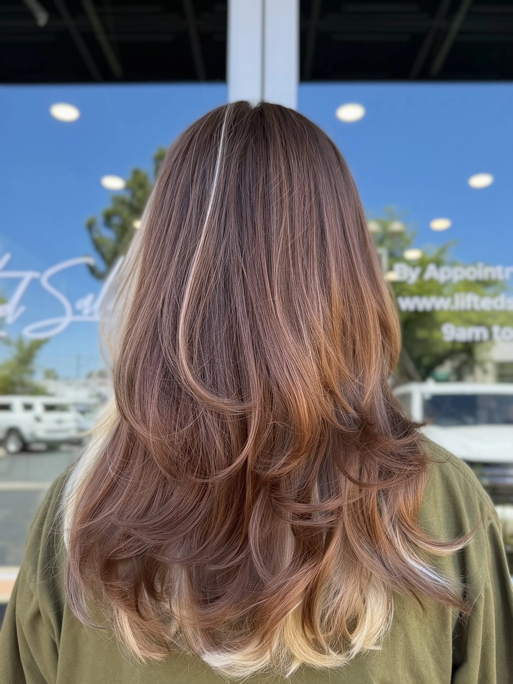 Back of a woman's head with long, wavy hair dyed in shades of pink, purple, and blonde, standing outside near a storefront.