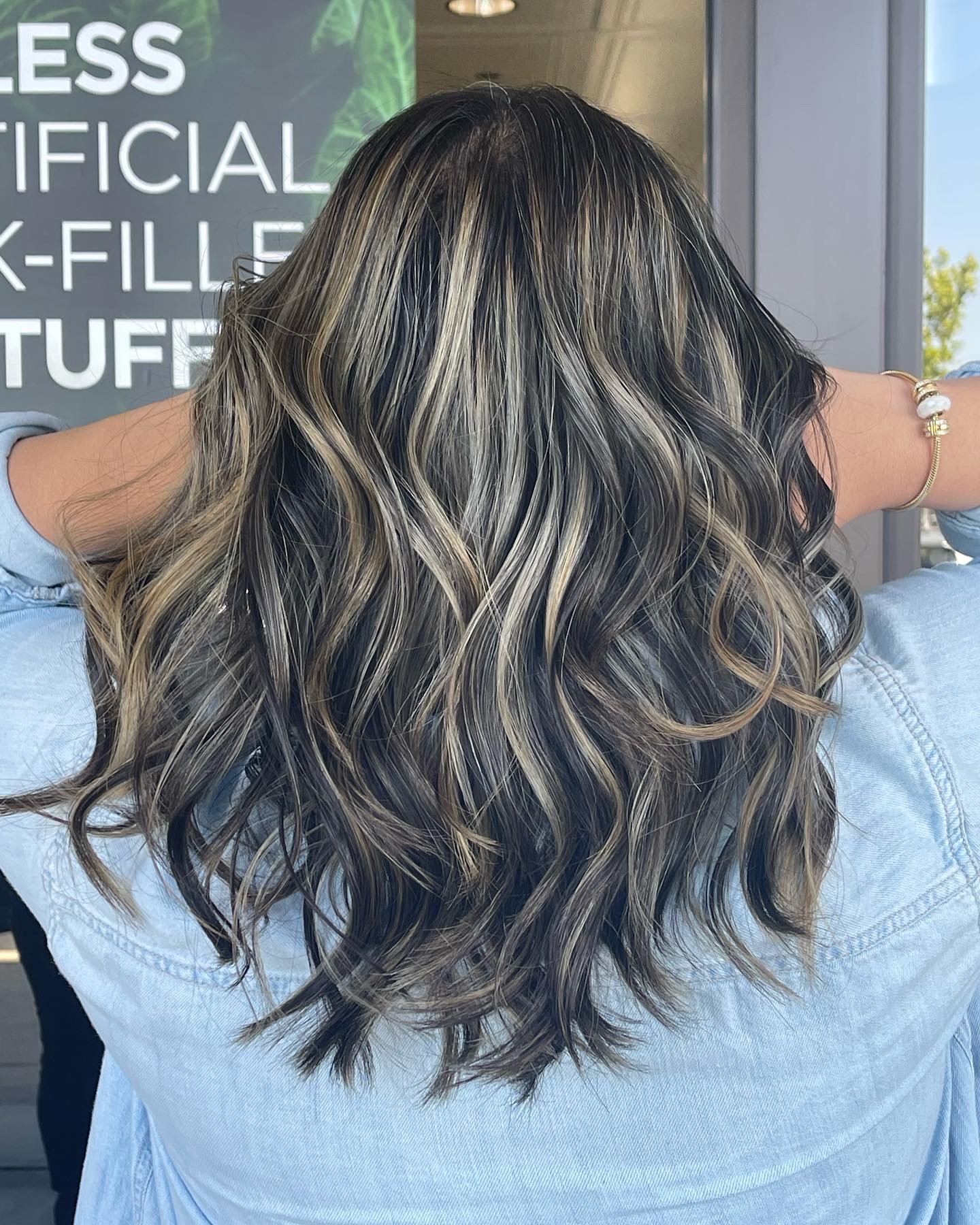 Back of woman with long, wavy, blonde and black highlighted hair, wearing a light blue denim shirt and a bracelet, standing near a window and a sign that reads 'LESS OFFICIAL PACK-FILLE... STUFF'.
