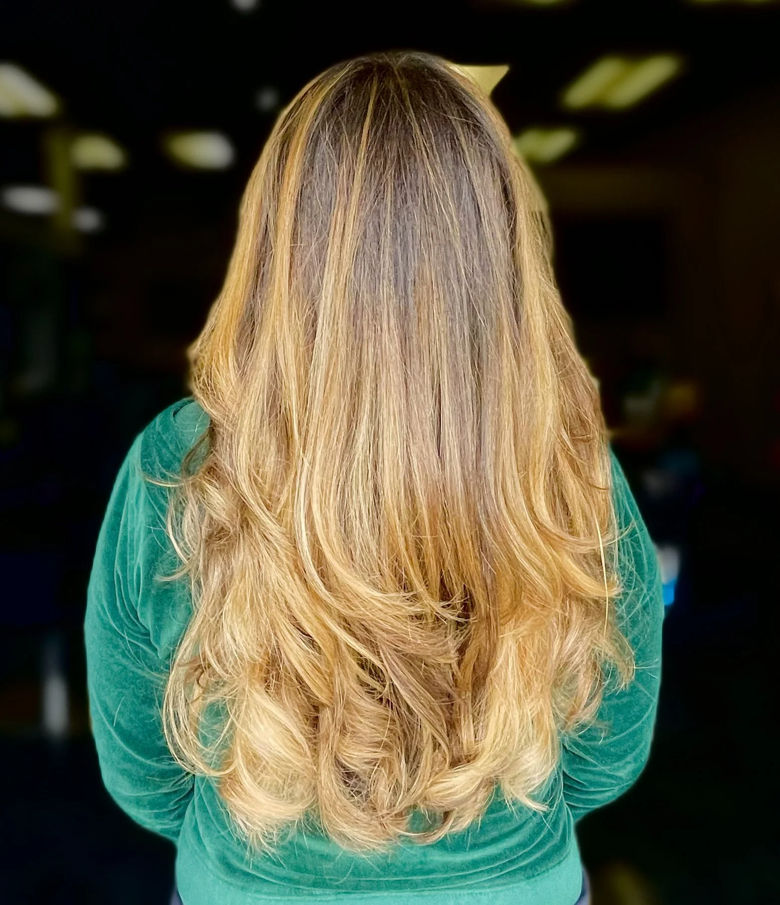 Back view of a woman with long, wavy blonde hair, wearing a teal long-sleeve shirt, in an indoor setting with blurred lighting.