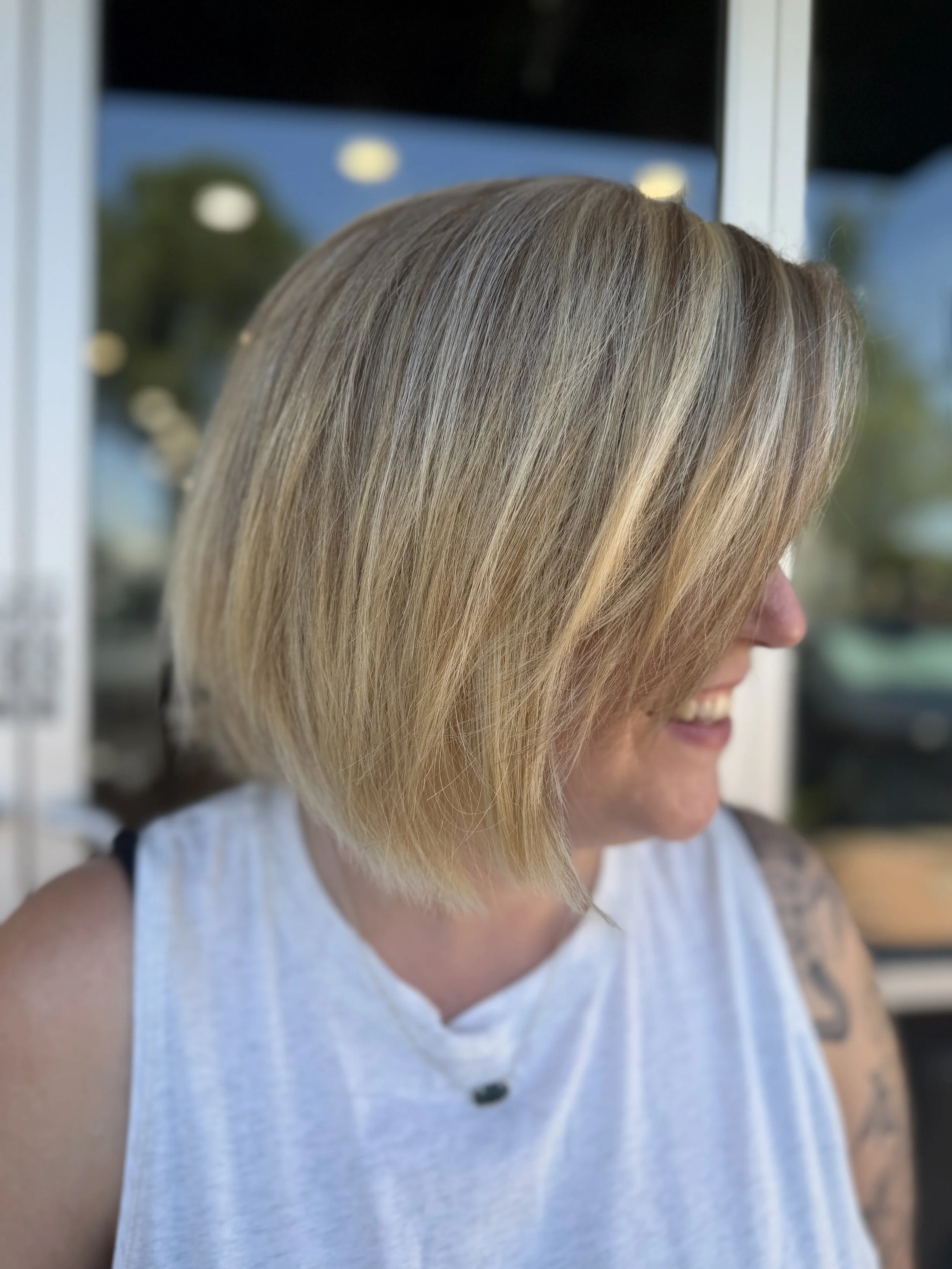 Close-up of a woman with shoulder-length highlighted ash cool blonde hair, wearing a sleeveless white shirt, smiling and looking down, with a blurred background of greenery and building.