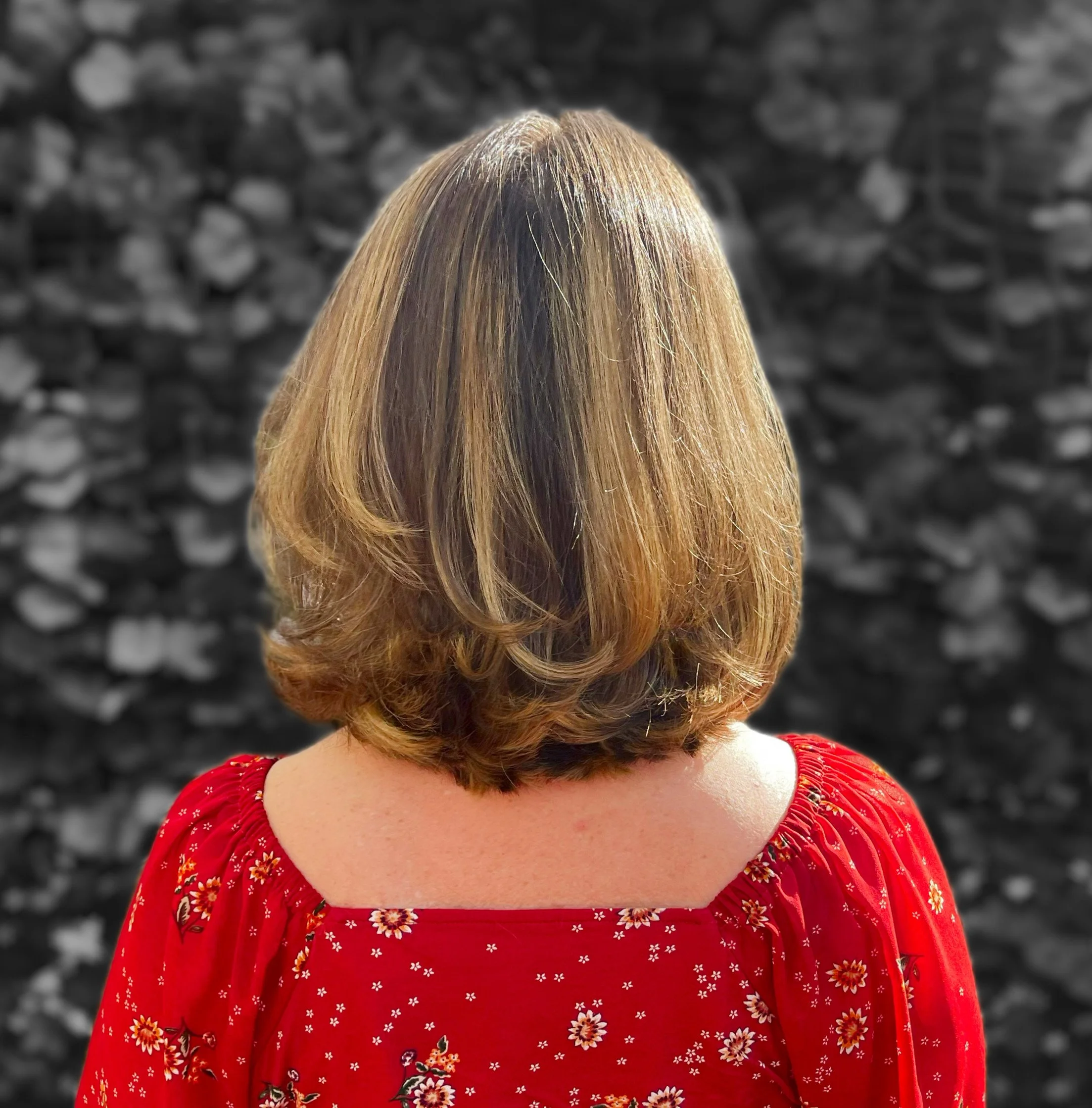 Back view of a woman with shoulder-length, layered, light brown hair, wearing a red blouse with floral patterns, standing in front of a blurred black-and-white stone wall background.
