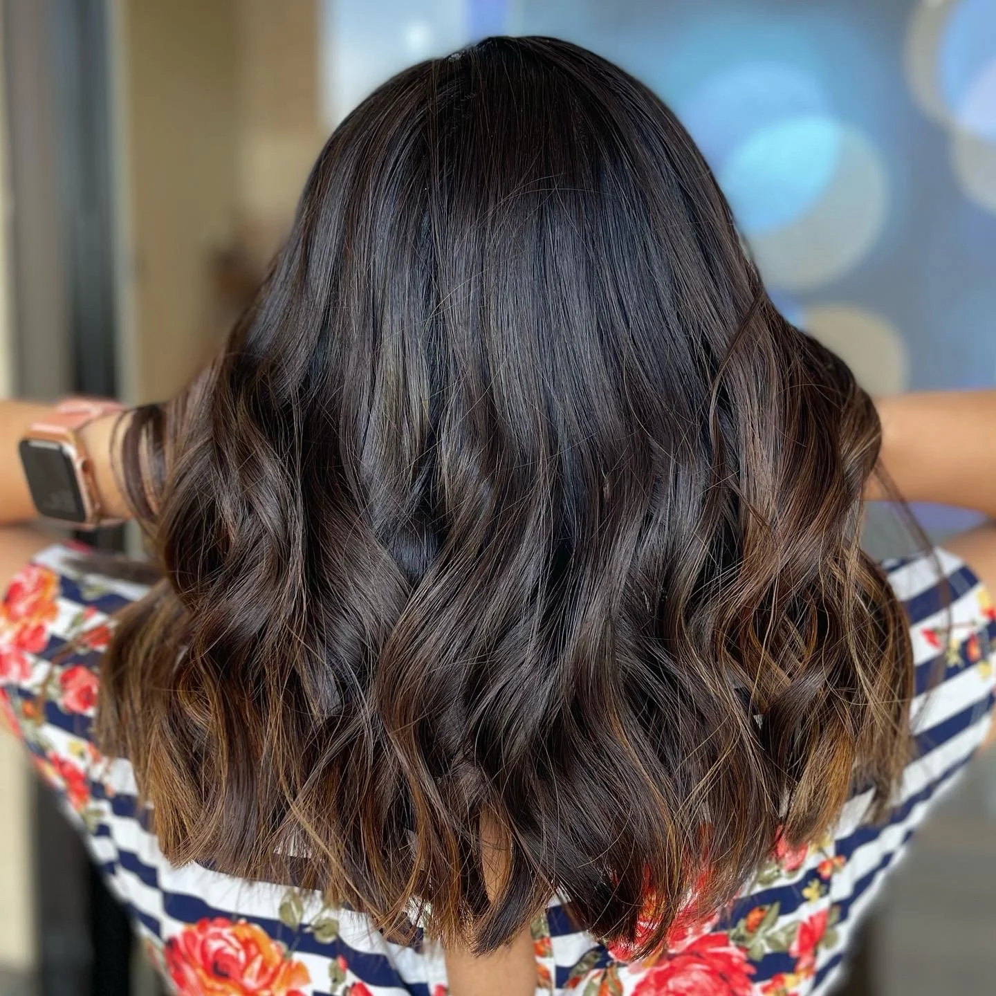 Back view of a woman with shoulder-length, dark brown, wavy hair wearing a floral top and a smartwatch.