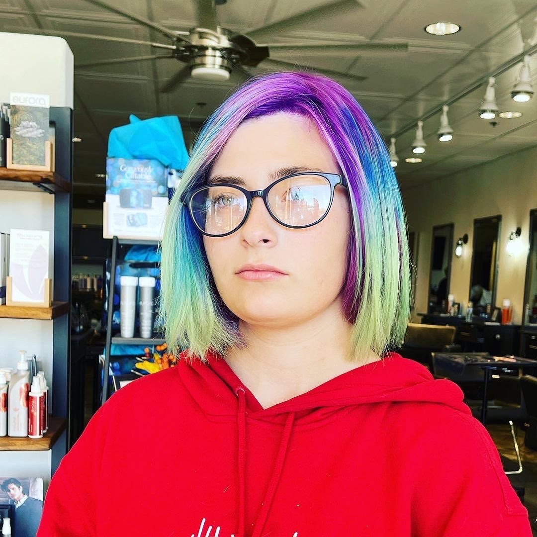Young woman with shoulder-length multi-colored hair in shades of purple, blue, and green, wearing glasses and a red hoodie, inside a salon or barbershop with hair product shelves and mirrors in the background.