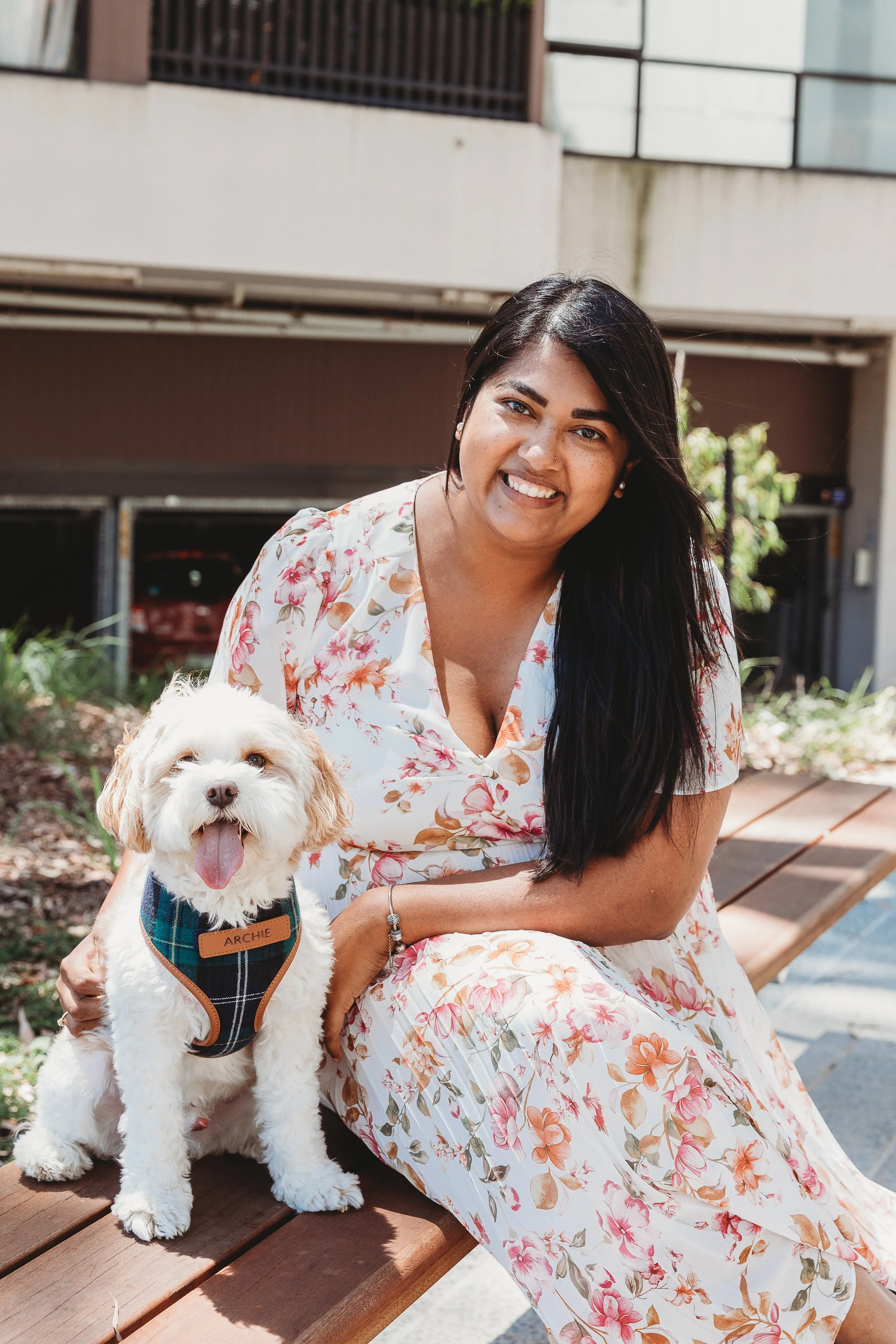A woman with long black hair wearing a floral dress sitting on a bench next to a small puppy with white and light brown fur, wearing a plaid harness, outdoors in a sunny setting.