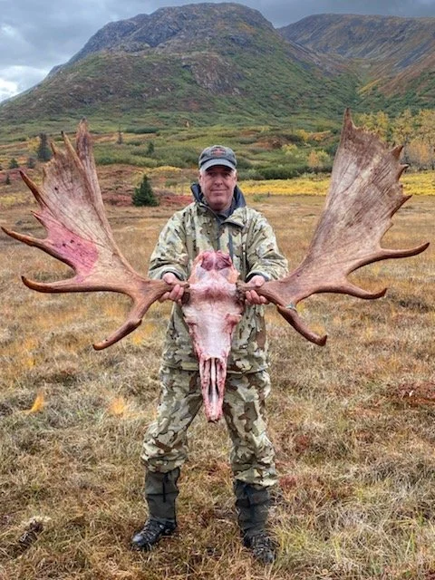 Person in camouflage clothing holding a large moose skull with antlers in a grassy field with mountains in the background.