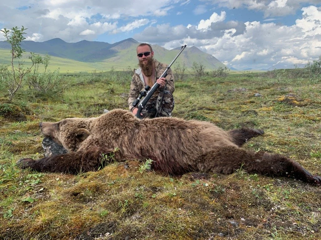 A man with a rifle, wearing sunglasses and camouflage clothing, kneeling behind a large, dead bear lying on the ground in an open, grassy landscape with mountains in the background.