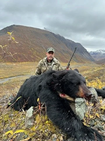 A man in camouflage clothing holding a rifle, kneeling behind a large dead black bear in an open landscape with mountains and cloudy sky.