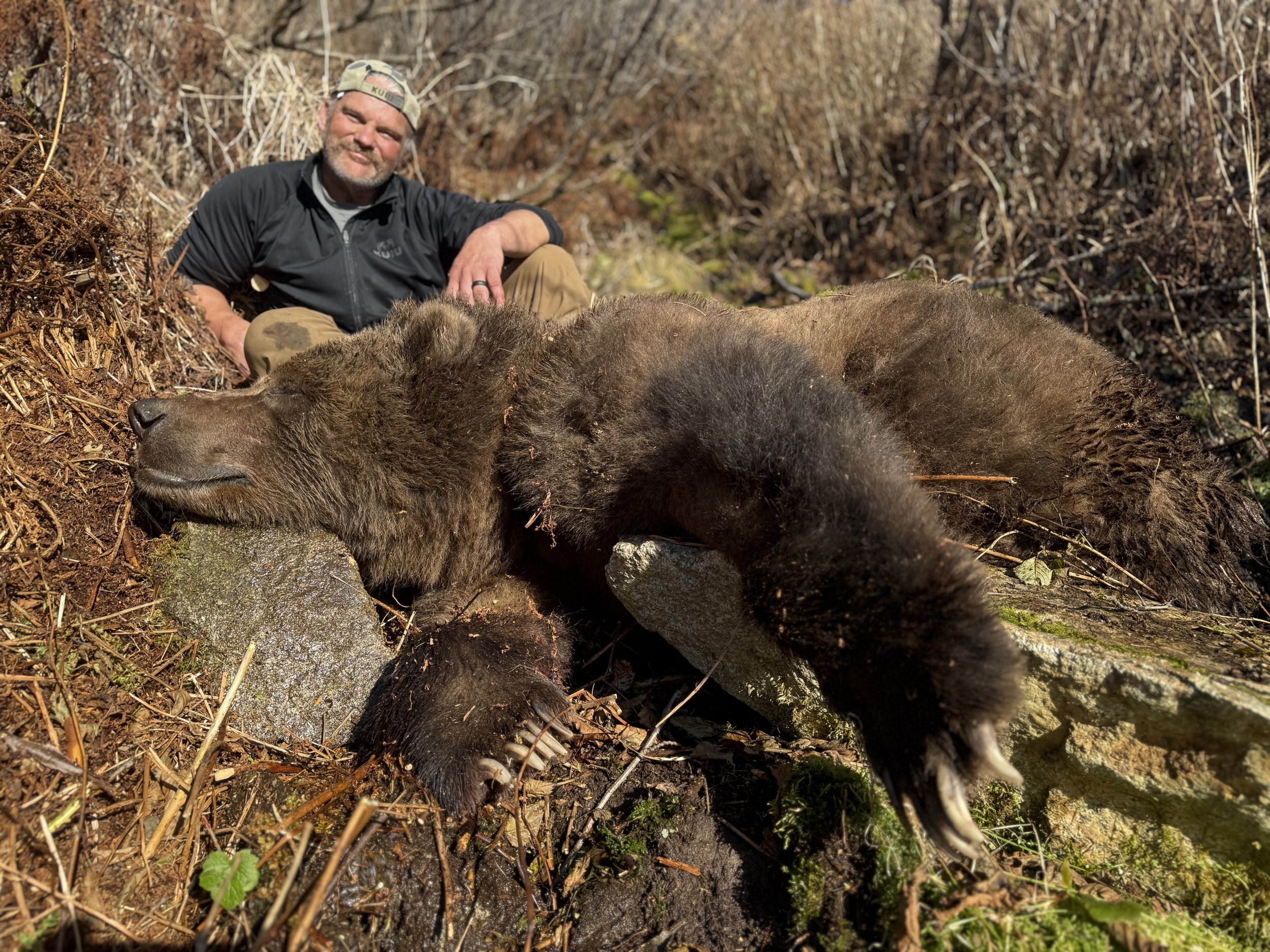 A man sitting on the ground outdoors, next to a dead bear, in a natural setting with dry bushes and rocks.