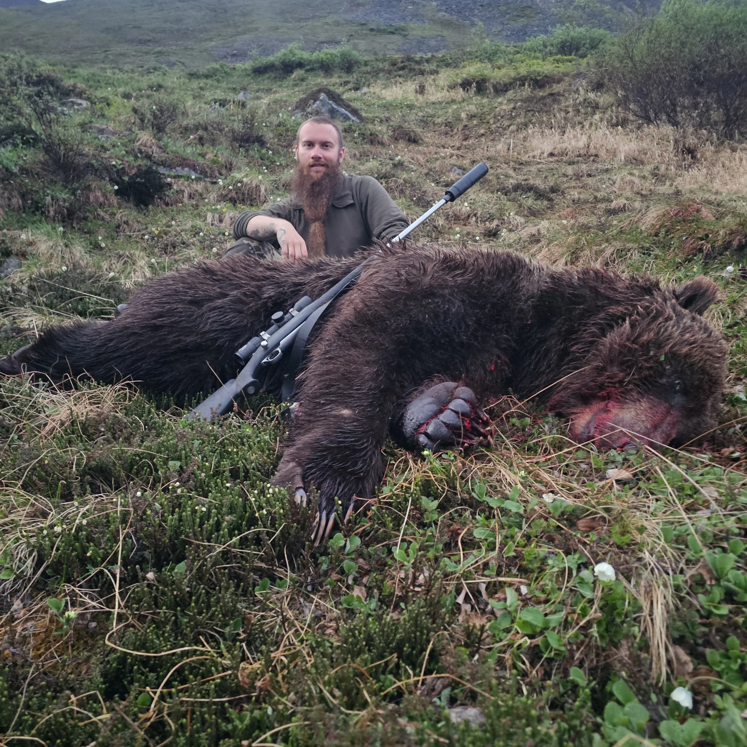 A man with a long beard sitting on grassy terrain in a remote natural setting, with lush green hills and shrubs in the background, holding a rifle.