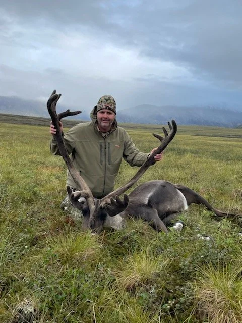 A man in outdoor gear kneels on the grass, holding the antlers of a large, deceased reindeer laying on the ground in a vast open field with mountains in the distance.