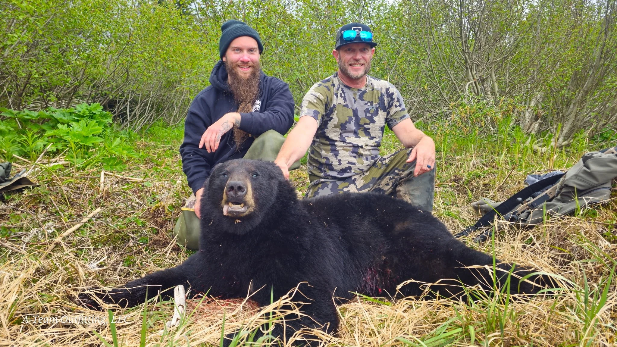 Two men kneeling behind a large black bear in a forested area, with green foliage and bushes around them. One man is wearing a black hoodie and beanie, the other is wearing a camouflage t-shirt and sunglasses. They are smiling and posing with the bear, which is lying on the ground with its mouth slightly open.