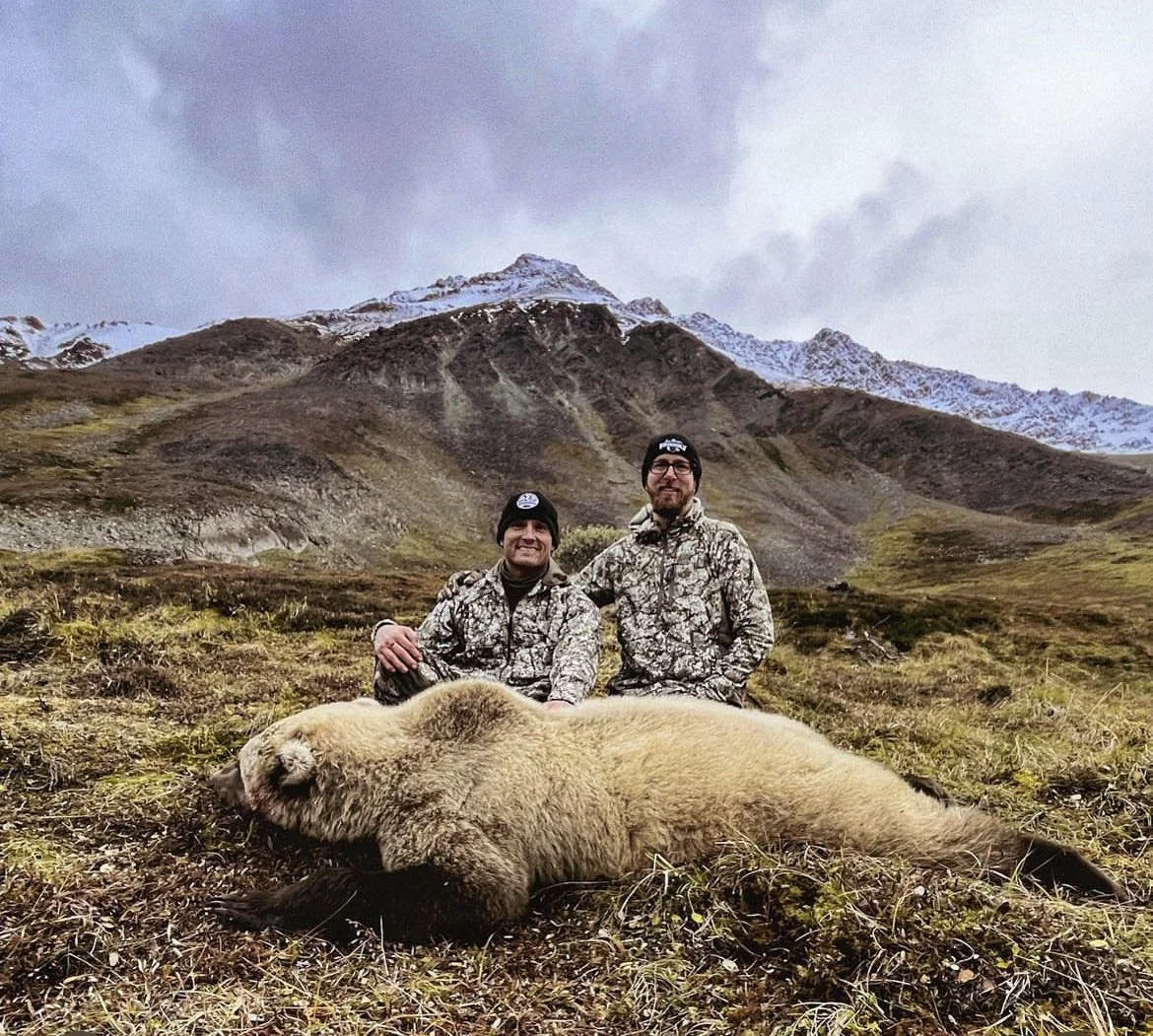 Two hunters in camouflage jackets and hats pose behind a large, dead bear lying on the ground in a mountainous landscape with snow-capped peaks in the background.