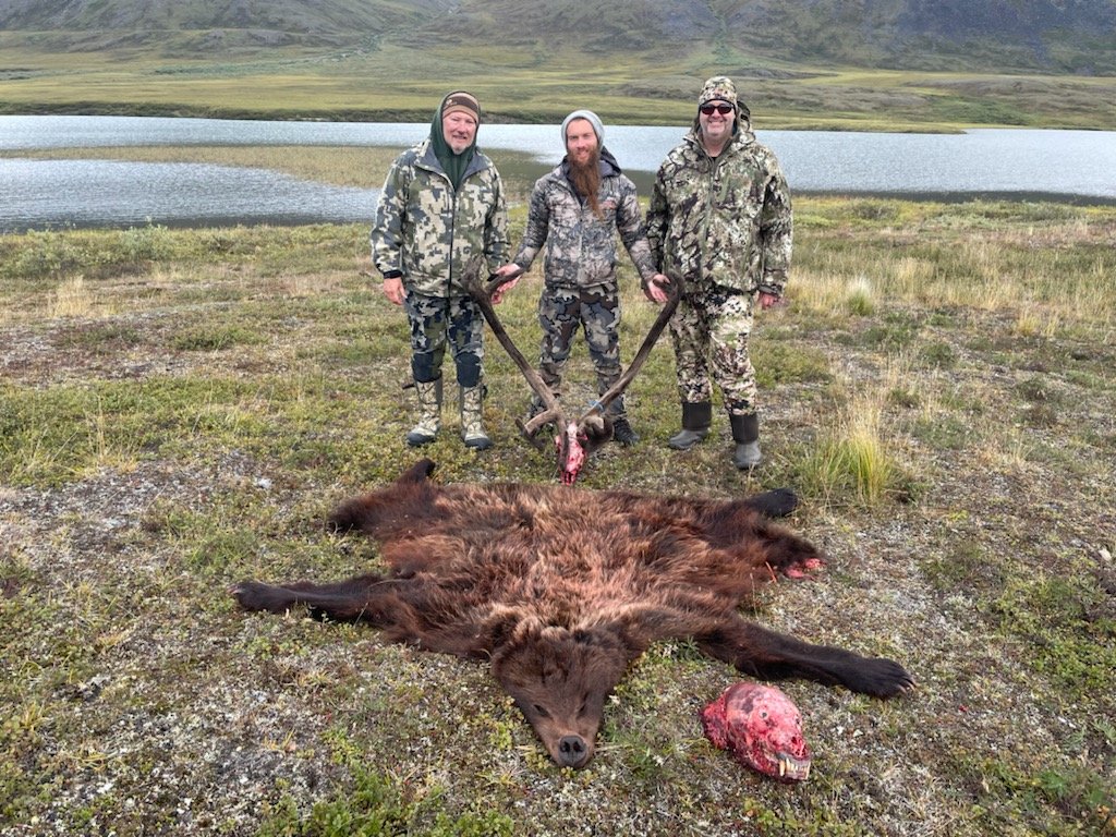 Three men in camouflage clothing posing with a dead bear lying on the ground in front of them, holding its antlers and a bear skull with exposed teeth, in a grassy landscape near a lake.