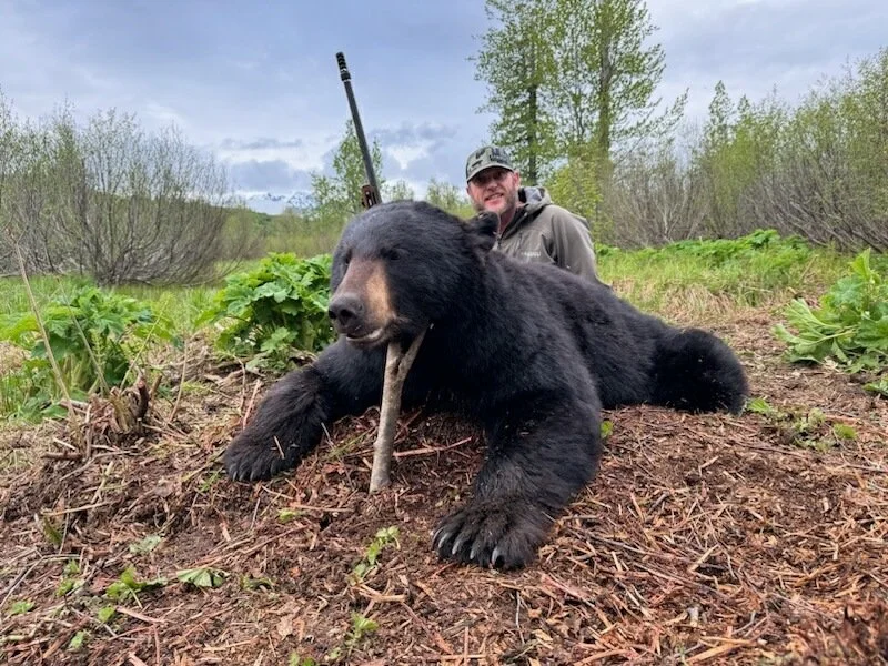A man in outdoor gear posing behind a large, dead bear lying on the ground in a green, wooded area.