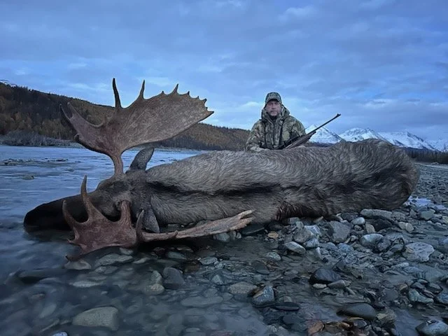 Hunter kneeling on rocky riverbank next to a large deceased moose with prominent antlers, in a mountainous landscape with snowy peaks in the background.