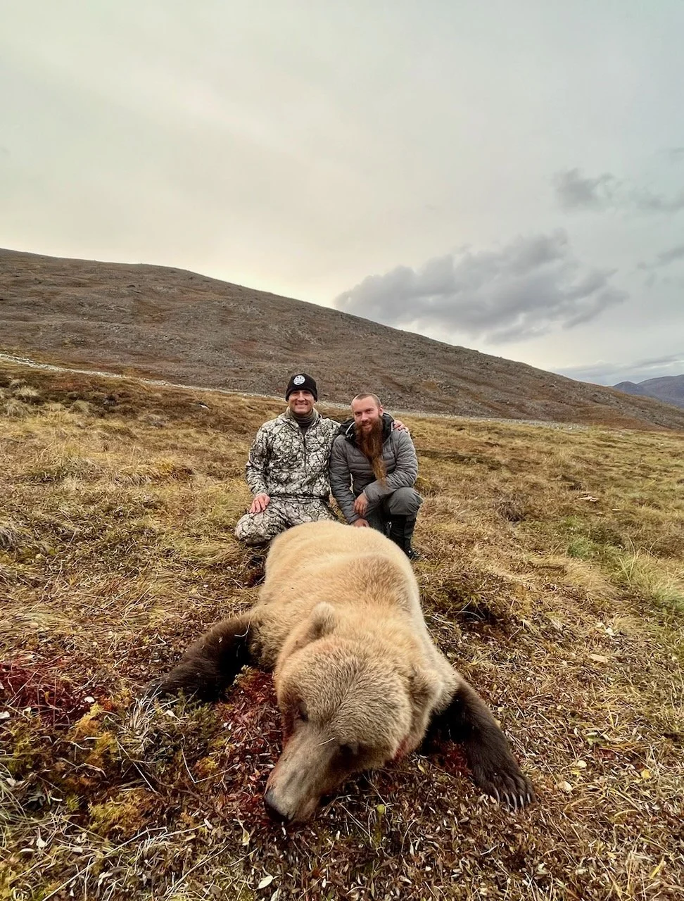 Two men kneeling in a grassy field with a large bear lying dead between them in a remote, mountainous landscape under a cloudy sky.