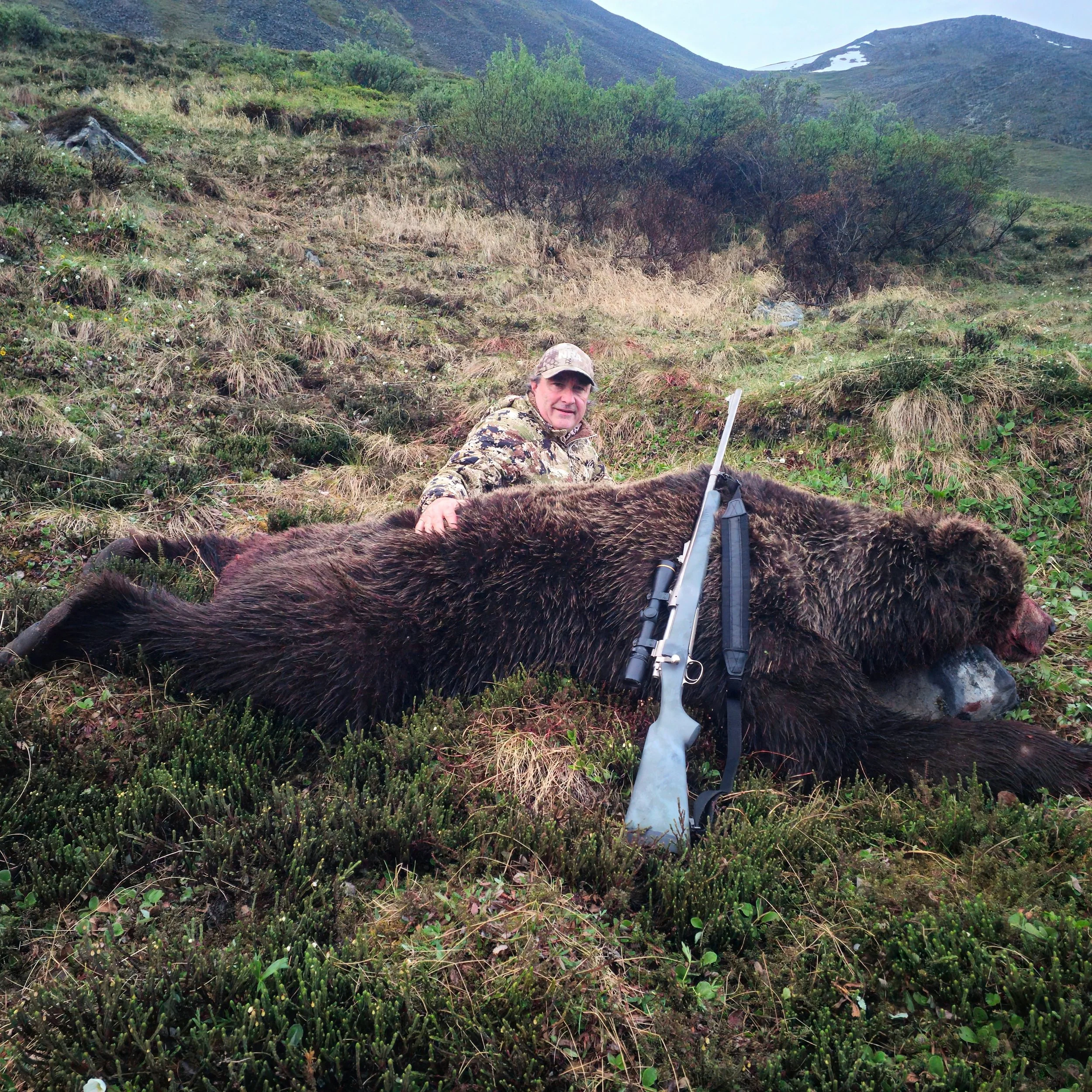 Man in camouflage clothing lying on the ground with a rifle, surrounded by greenery and mountain landscape.