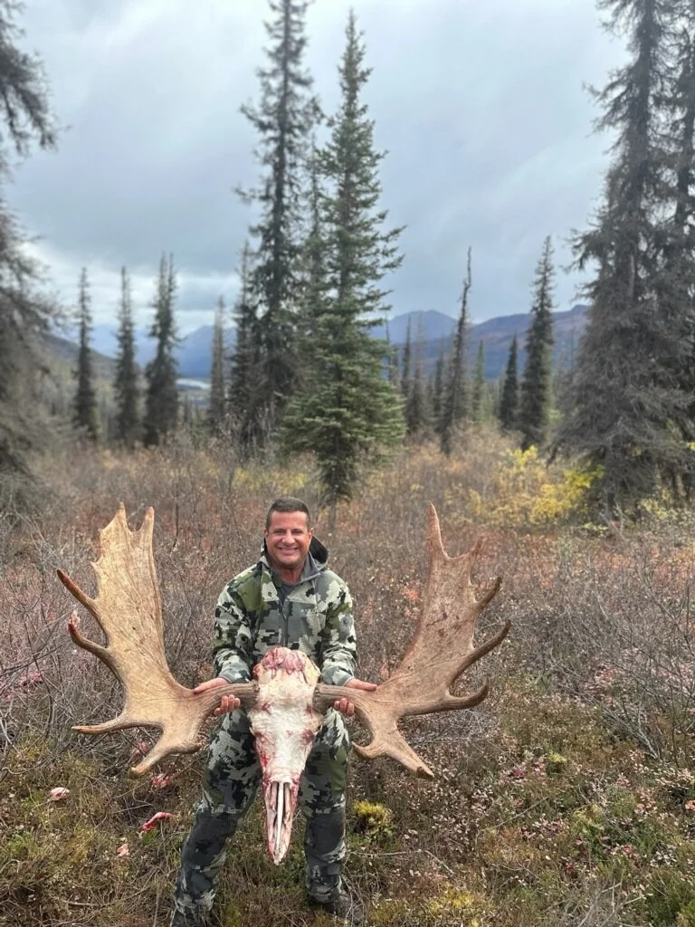 A man in camouflage clothing holding a large moose skull with antlers in a forest with trees and mountains in the background.