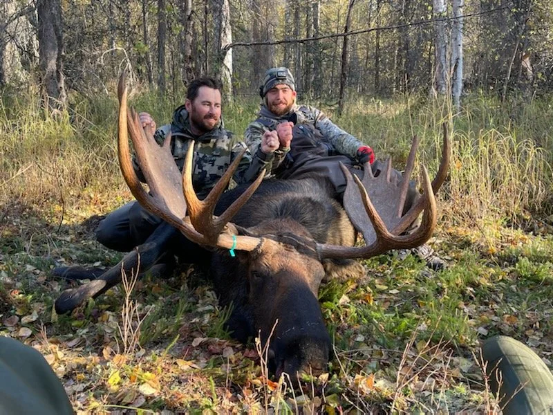 Two men sitting behind a large moose with impressive antlers in a forested area, celebrating with clenched fists.