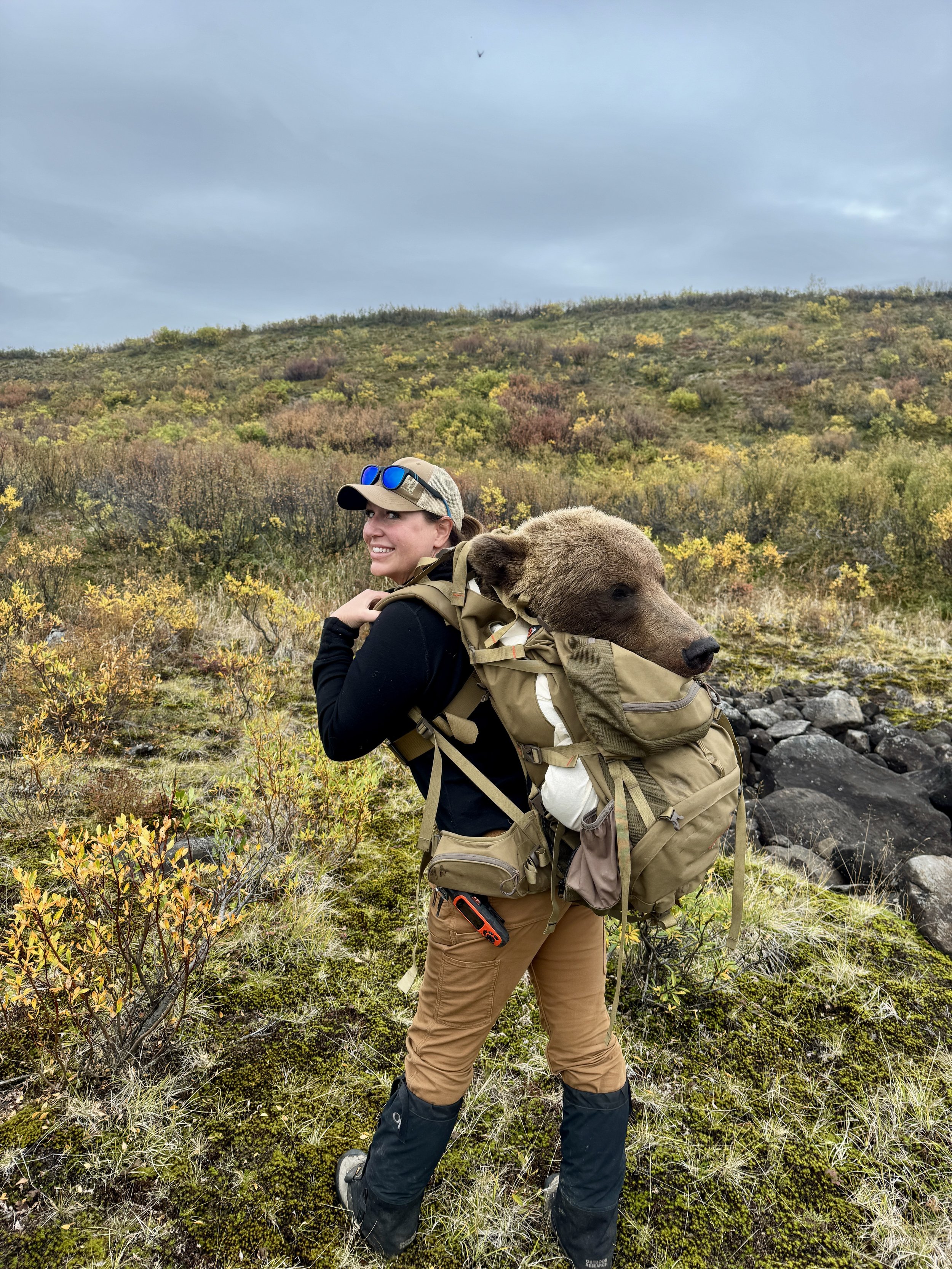 A woman in outdoor hiking gear carrying a bear-shaped backpack in a natural landscape with bushes and a hillside, under a cloudy sky.