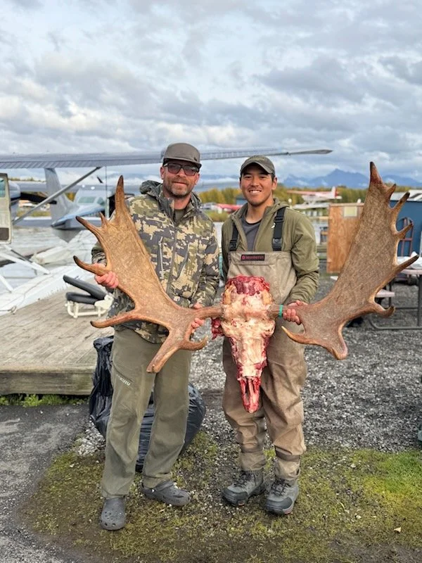 Two men standing outdoors holding a large moose skull with antlers. They are dressed in outdoor gear, and there are airplanes and a cloudy sky in the background.