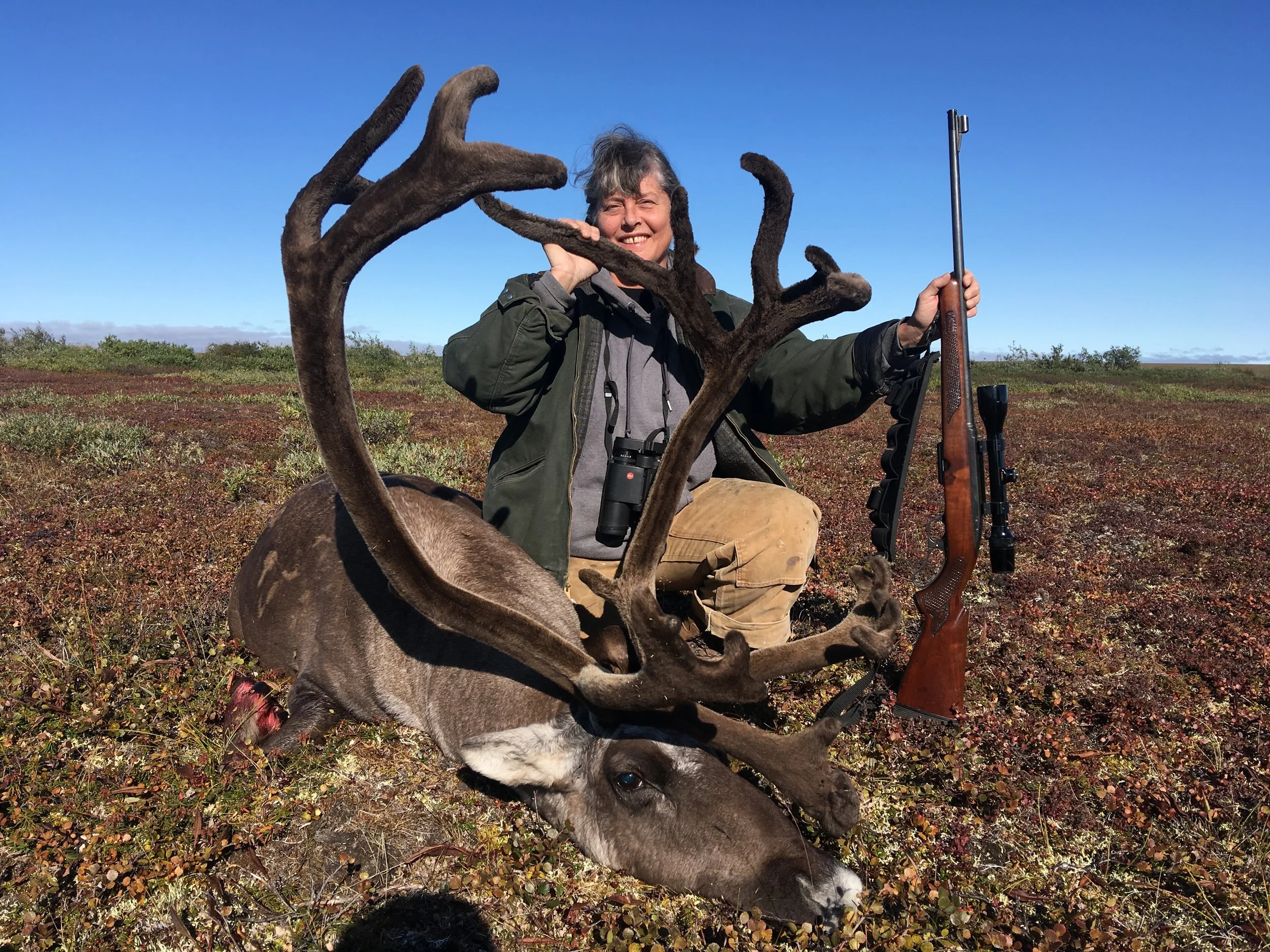 A person in outdoor clothing holding a rifle, posing with a reindeer in a field with a clear blue sky. The person is smiling and wearing binoculars around their neck.