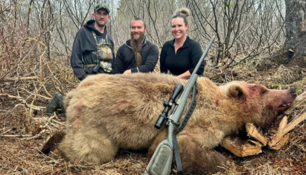 Three smiling hunters standing behind a dead bear with a rifle resting on the bear's back, in a wooded area.
