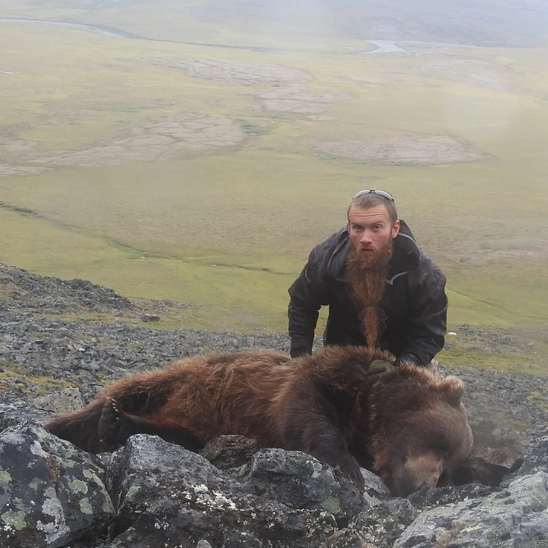 A man with a long beard and a black jacket is kneeling on a rocky hillside next to a large, brown bear lying on the rocks with its head resting on the ground, in a green, mountainous landscape.
