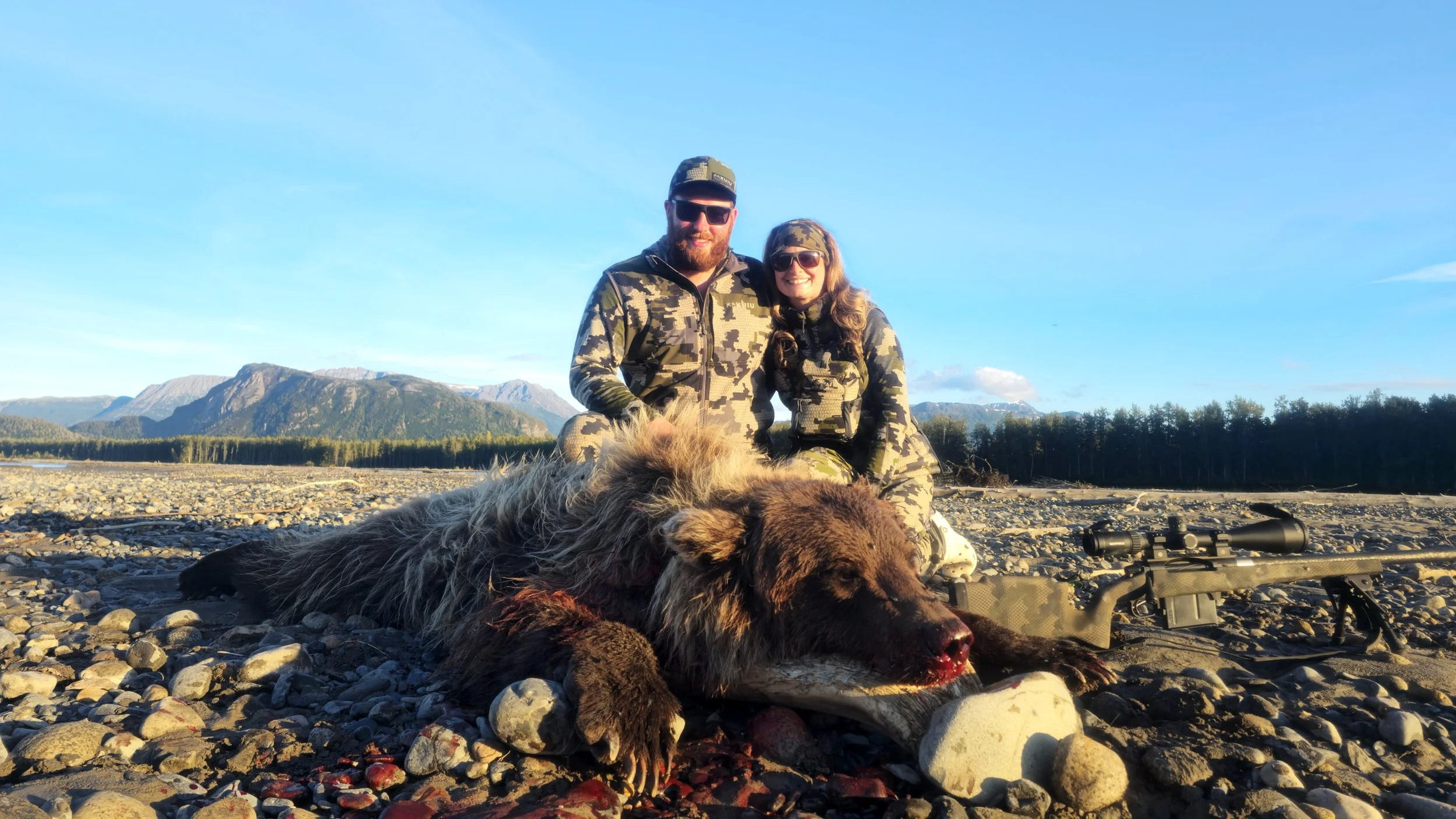 Two people in camouflage clothing kneel beside a large dead bear on a rocky outdoor landscape with mountains and a blue sky, holding a rifle.