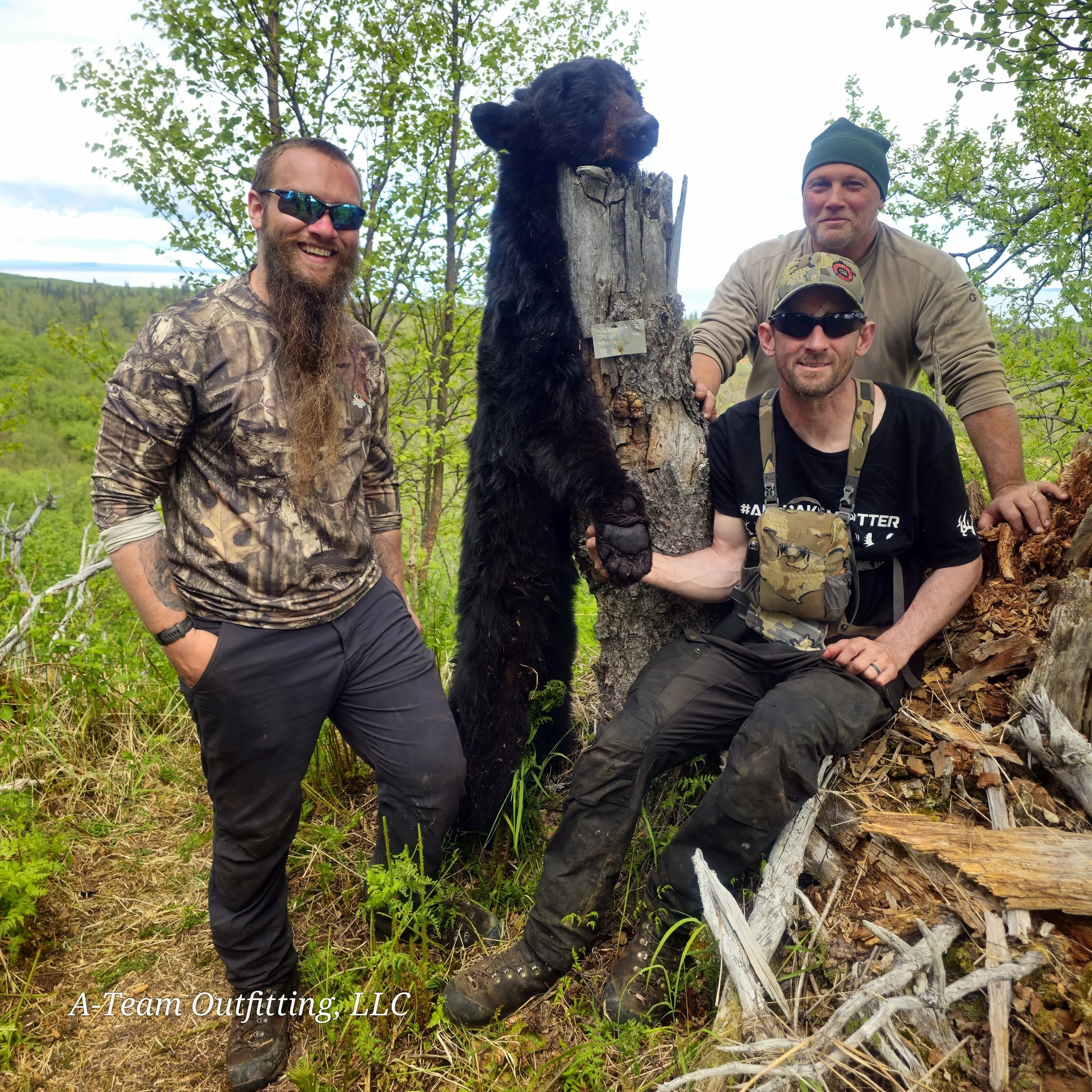 Three men posing outdoors in a forested area, with one man sitting on a log and wearing sunglasses, another standing with a hat and sunglasses, and a third man with a long beard and camouflage clothing. A black bear mount is mounted on a tree stump between them.