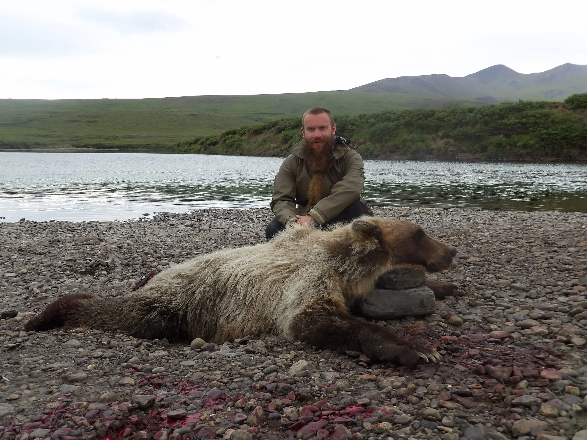 A bearded man in outdoor clothing sitting on a rocky shoreline near a body of water with green hills and mountains in the background.