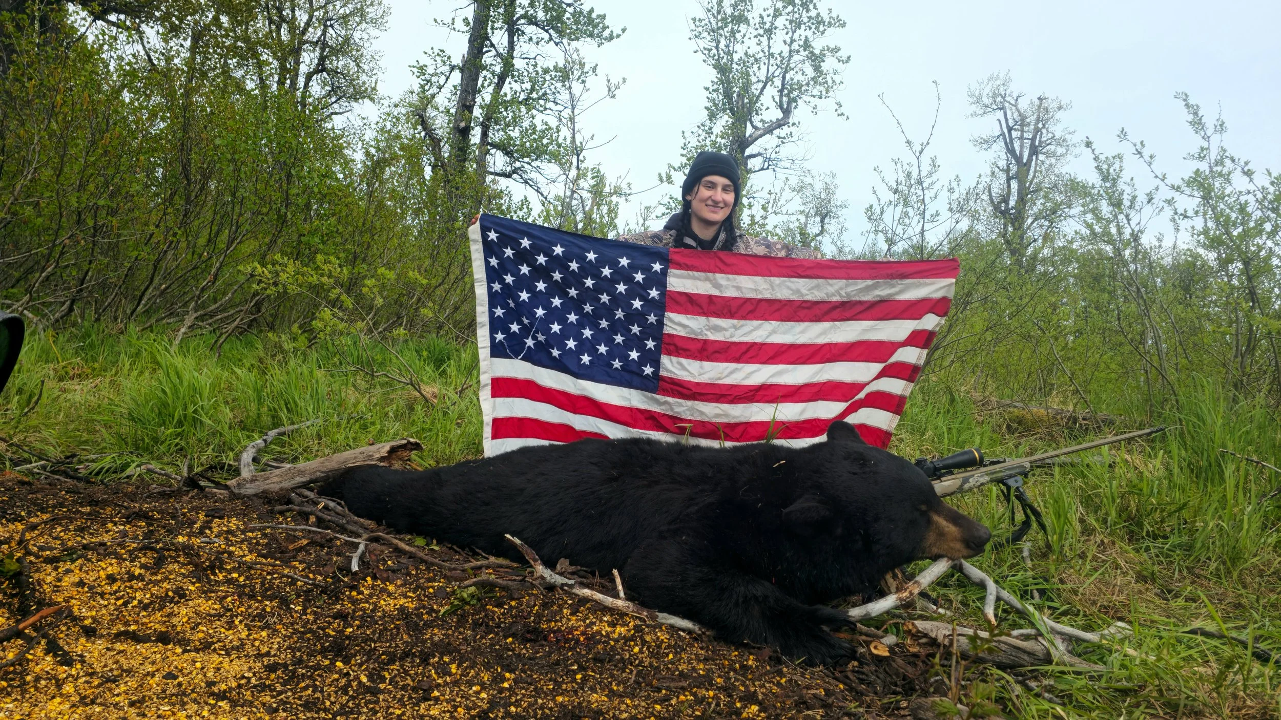 A young woman with dark hair in a black beanie, smiling and holding an American flag, stands in a forested area with green trees and grass. A dead black bear lies on the ground in front of her, with a rifle resting against it. The ground is covered with yellow and orange debris.