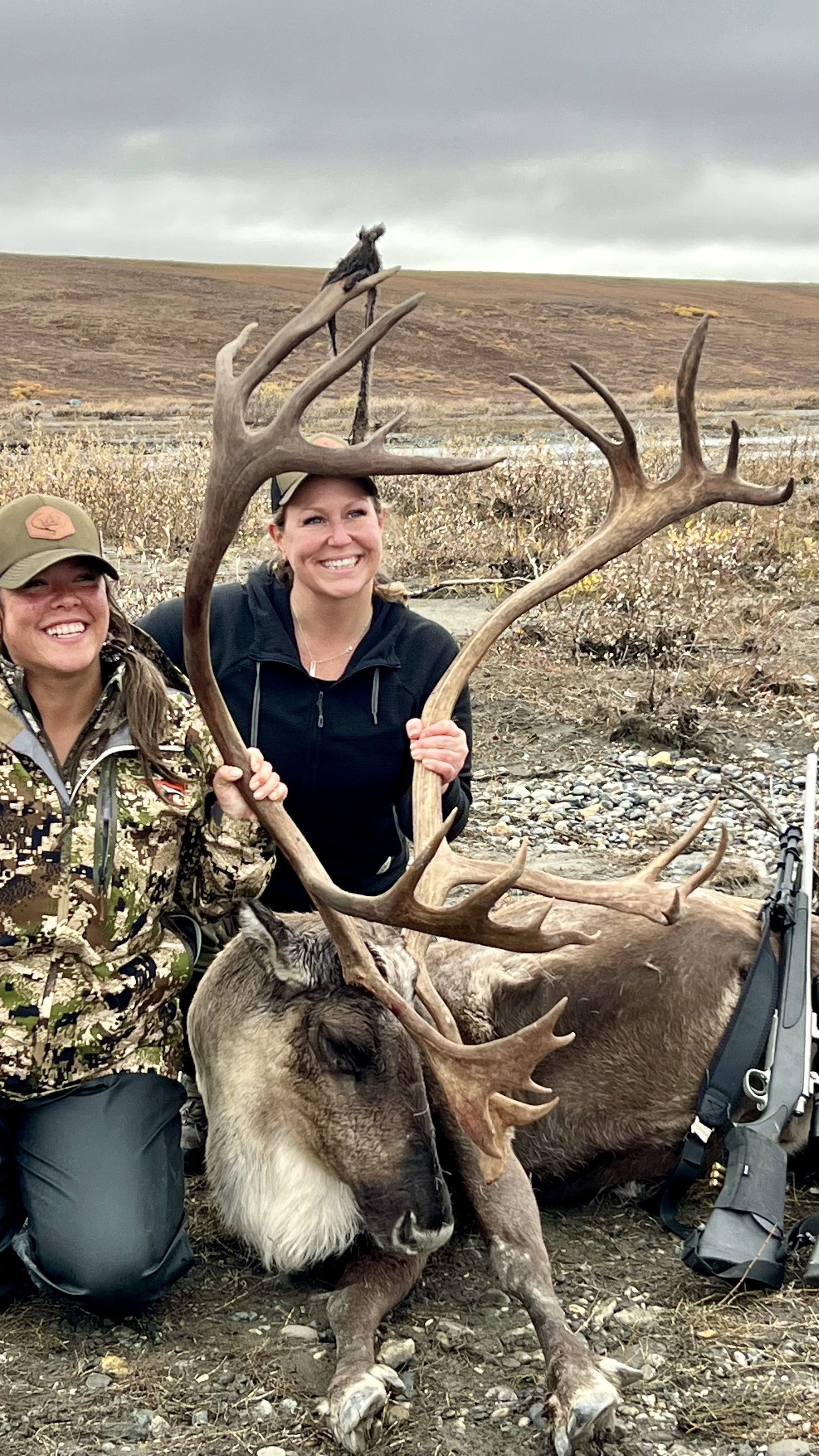 Two women in outdoor gear smiling and posing with a large moose they have hunted, in a rugged outdoor landscape with a cloudy sky.