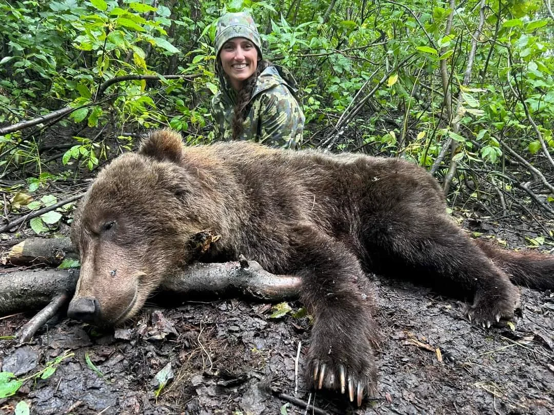 A smiling woman in camouflage clothes and a hat crouching behind a large, lifeless bear lying on the muddy ground in a forest.