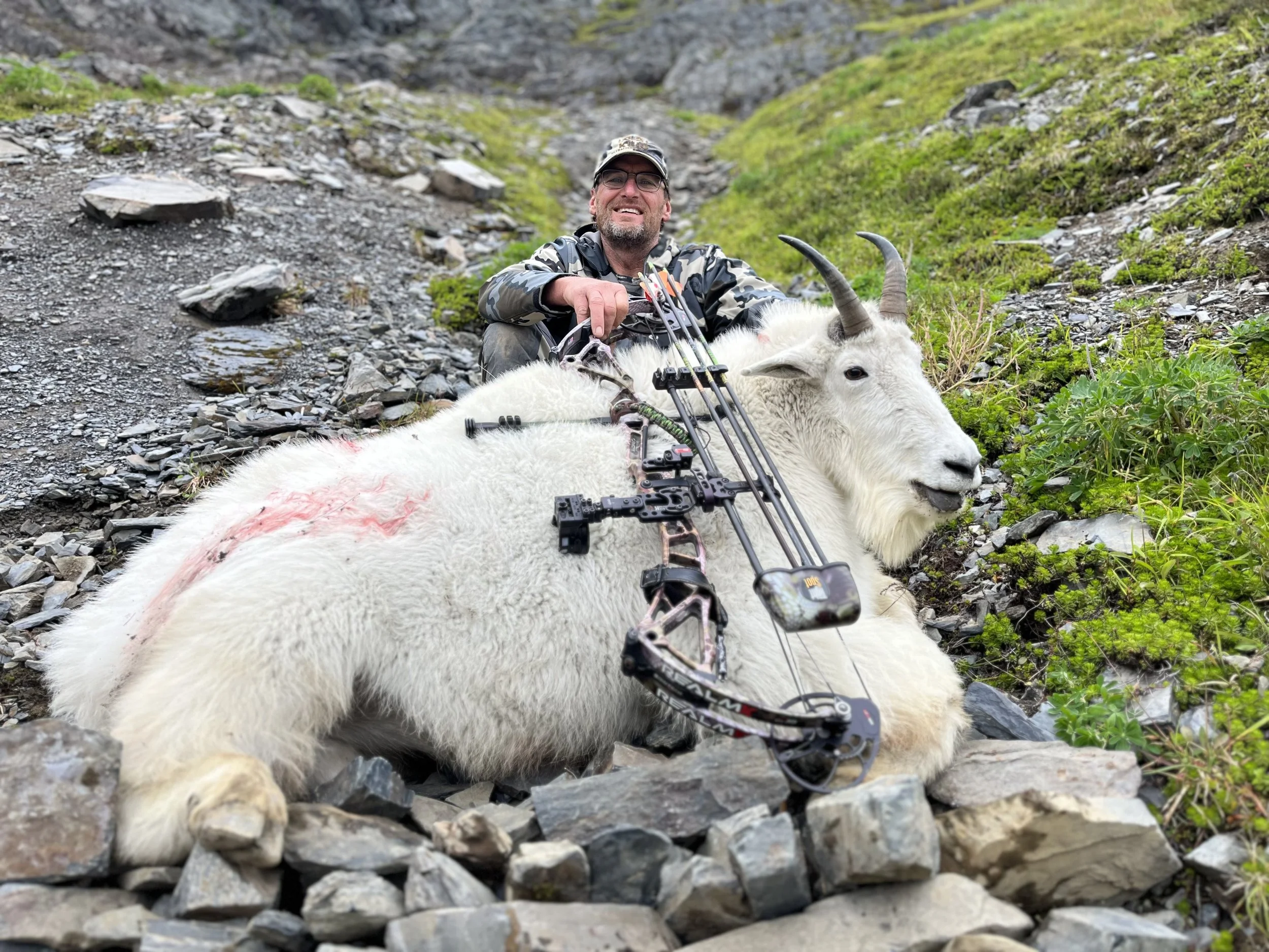 A man with glasses and a camouflage jacket sitting on rocky ground, smiling, holding a bow, beside a white mountain goat lying on the rocks.