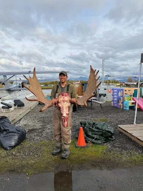 Man standing outdoors in a marina area holding a large moose skull with antlers, smiling, with planes and children’s play area in the background.