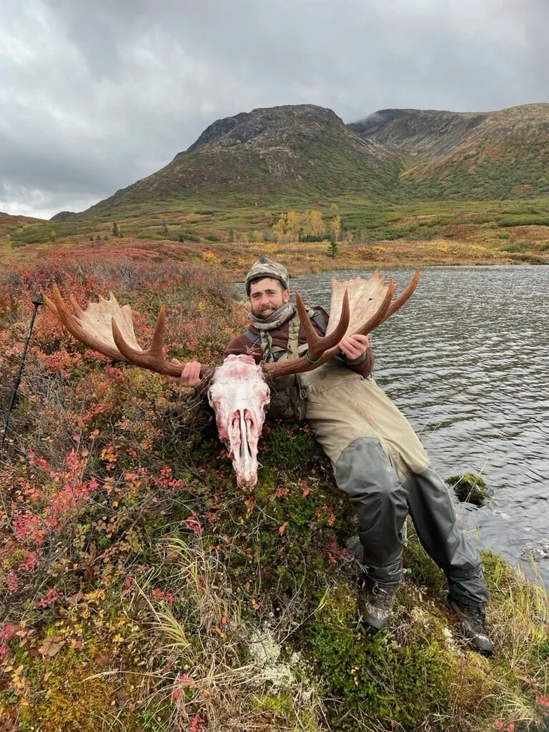 A man outdoors near a lake holding a moose skull and antlers, with colorful bushes and mountains in the background.