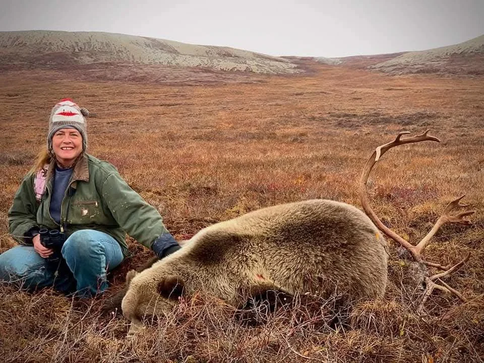 A woman in outdoor clothing, sitting on the ground, with a mountain landscape in the background, smiling next to a dead bear and a fallen elk with antlers.