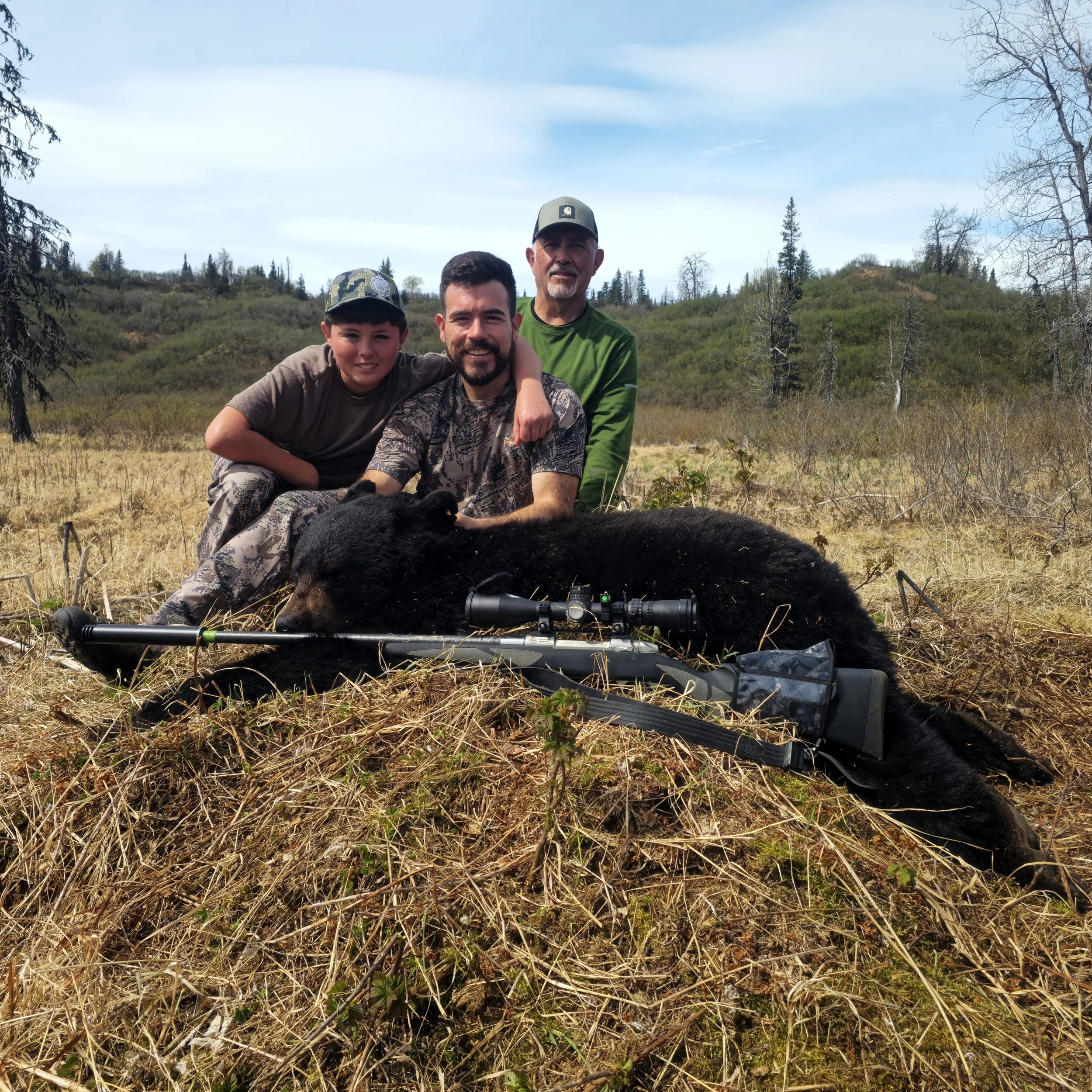 Three people, two men and a boy, posing outdoors with a bear they have hunted, with hunting gear including a rifle and scope in front of the bear, in a grassy, wooded landscape under a partly cloudy sky.