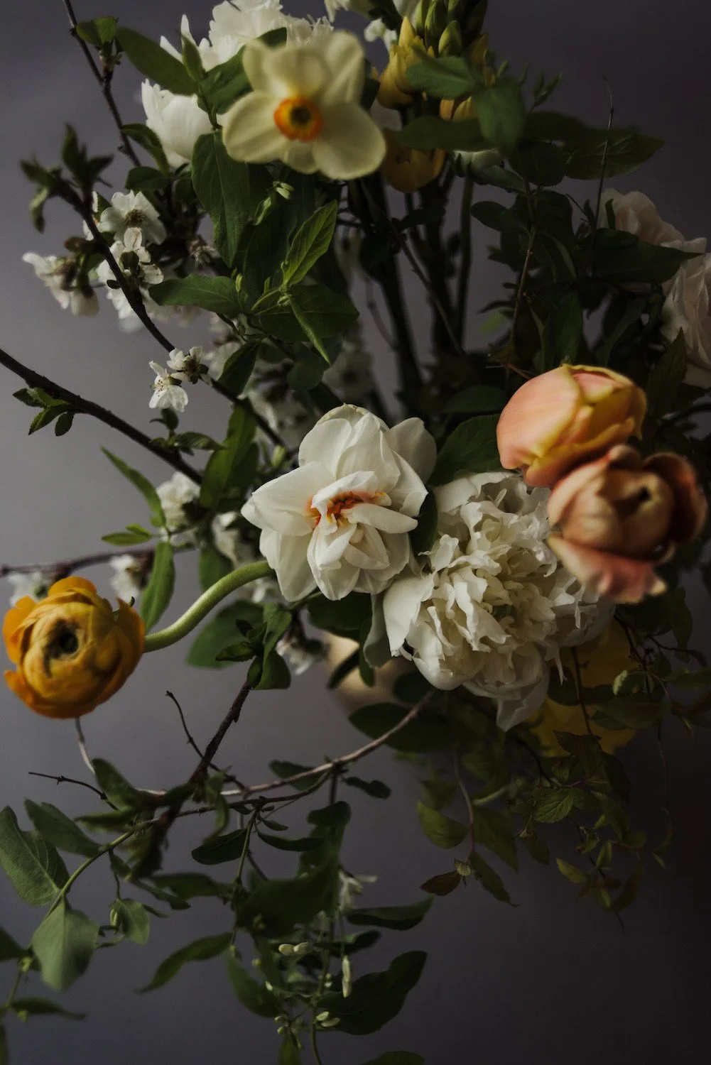 A bouquet of white, yellow, and orange flowers, including roses and peonies, with green leaves and dark stems against a dark background.