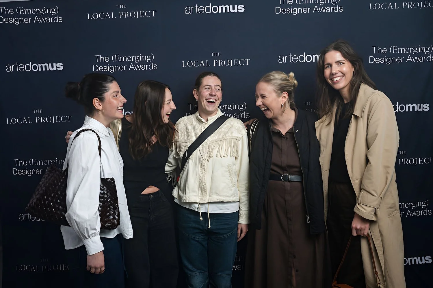Five women smiling and laughing together at an event with a dark blue backdrop featuring logos and text for 'The Emerging Designer Awards' and 'artedomeus'.