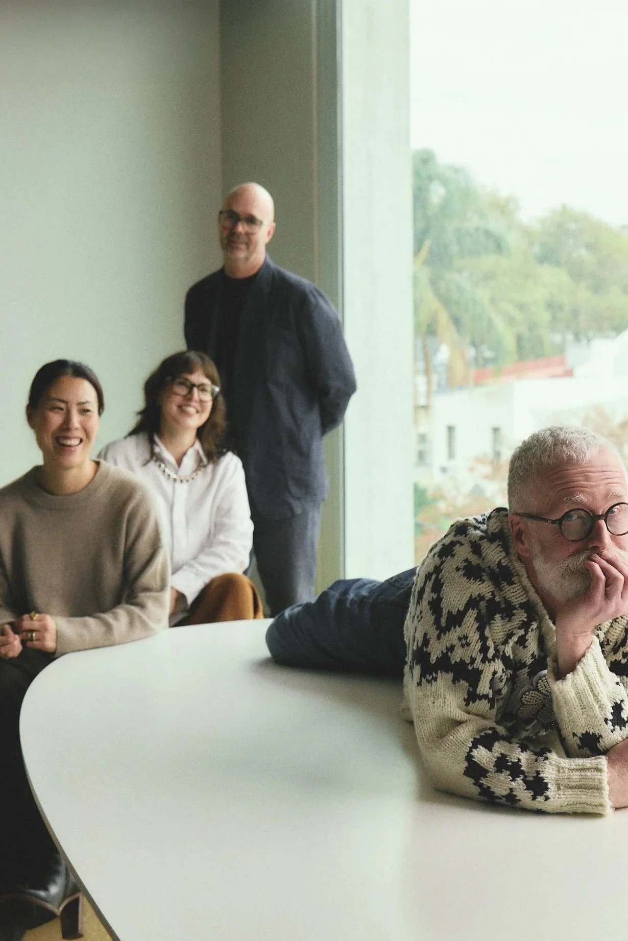 Four people in a meeting room near a large window with trees outside. Three are seated at a white table and one person is standing behind them. The seated group includes a woman with a beige sweater, a woman with glasses and a white jacket, and a man with glasses and a patterned sweater. The man standing is wearing a dark jacket and glasses.