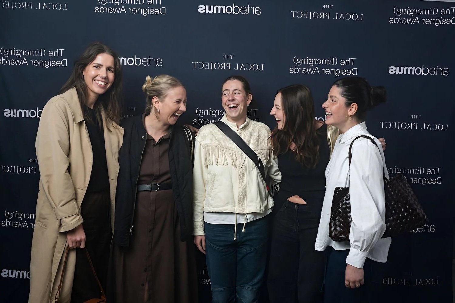 Group of five women standing together, smiling and laughing in front of a black backdrop with white text.