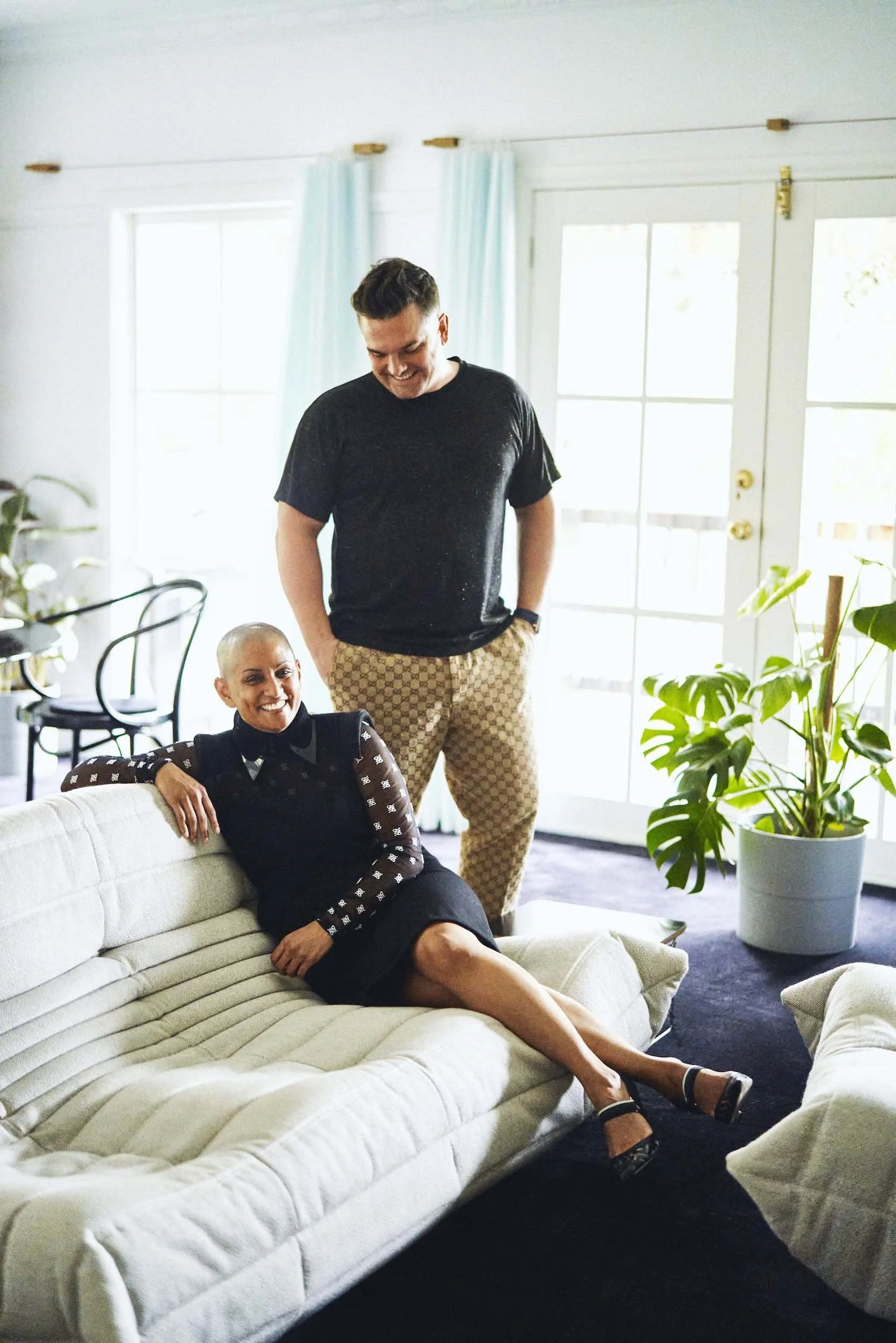 Max Soans Burne with a shaved head sitting on a white couch, smiling, wearing a black dress with white patterns, black heels, with Richard Burne standing in a bright living room with large windows and plants.