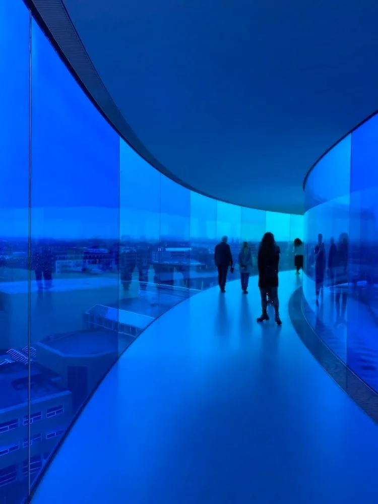 People walking on a curved glass corridor with blue-tinted glass walls and ceiling, overlooking a cityscape at dusk or night.