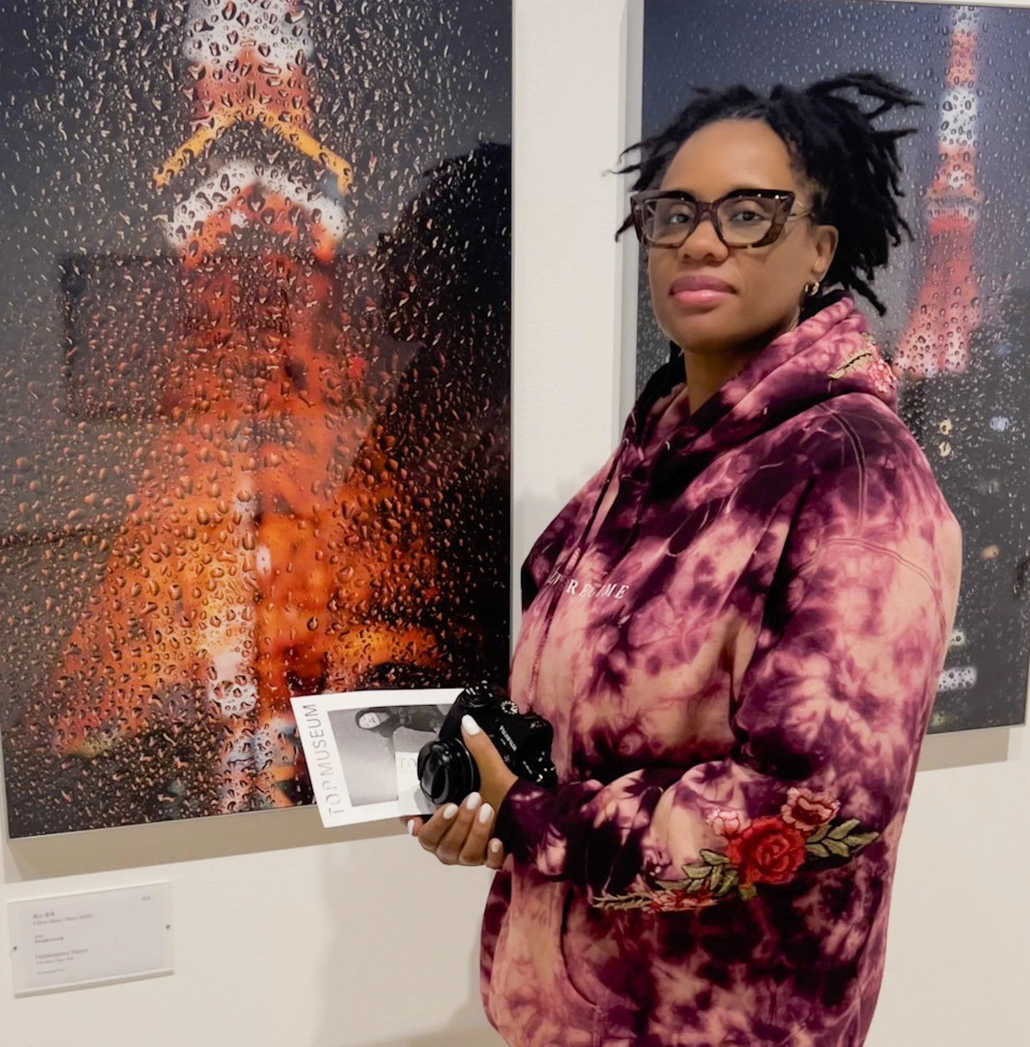 Woman with glasses and dreadlocks wearing a tie-dye hoodie featuring floral embroidery, holding a camera, standing in front of an art exhibit with a piece of artwork covered in raindrop-like patterns.