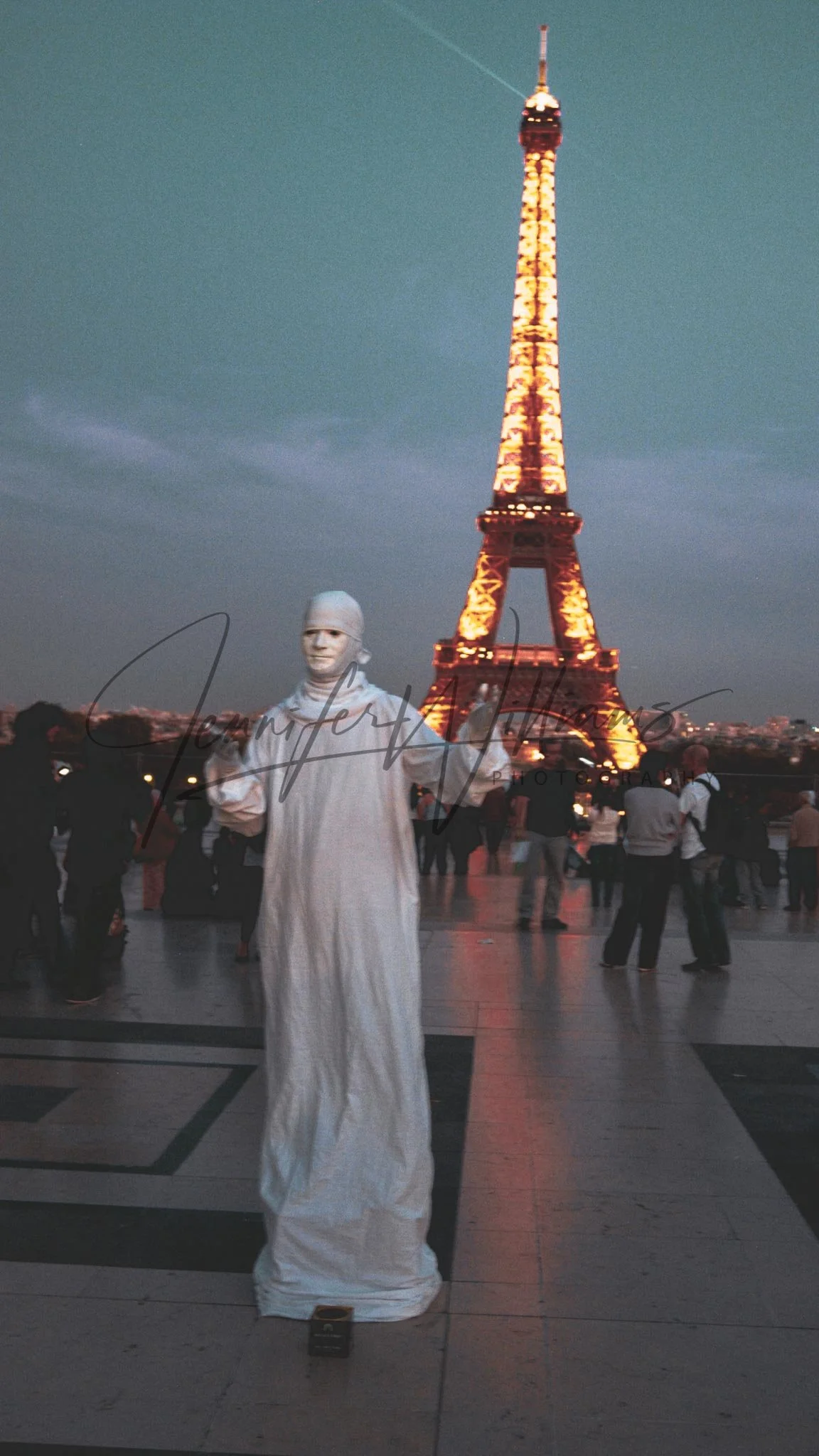 A person dressed as a living statue in white robes standing in front of the Eiffel Tower illuminated at night, surrounded by a crowd of people.