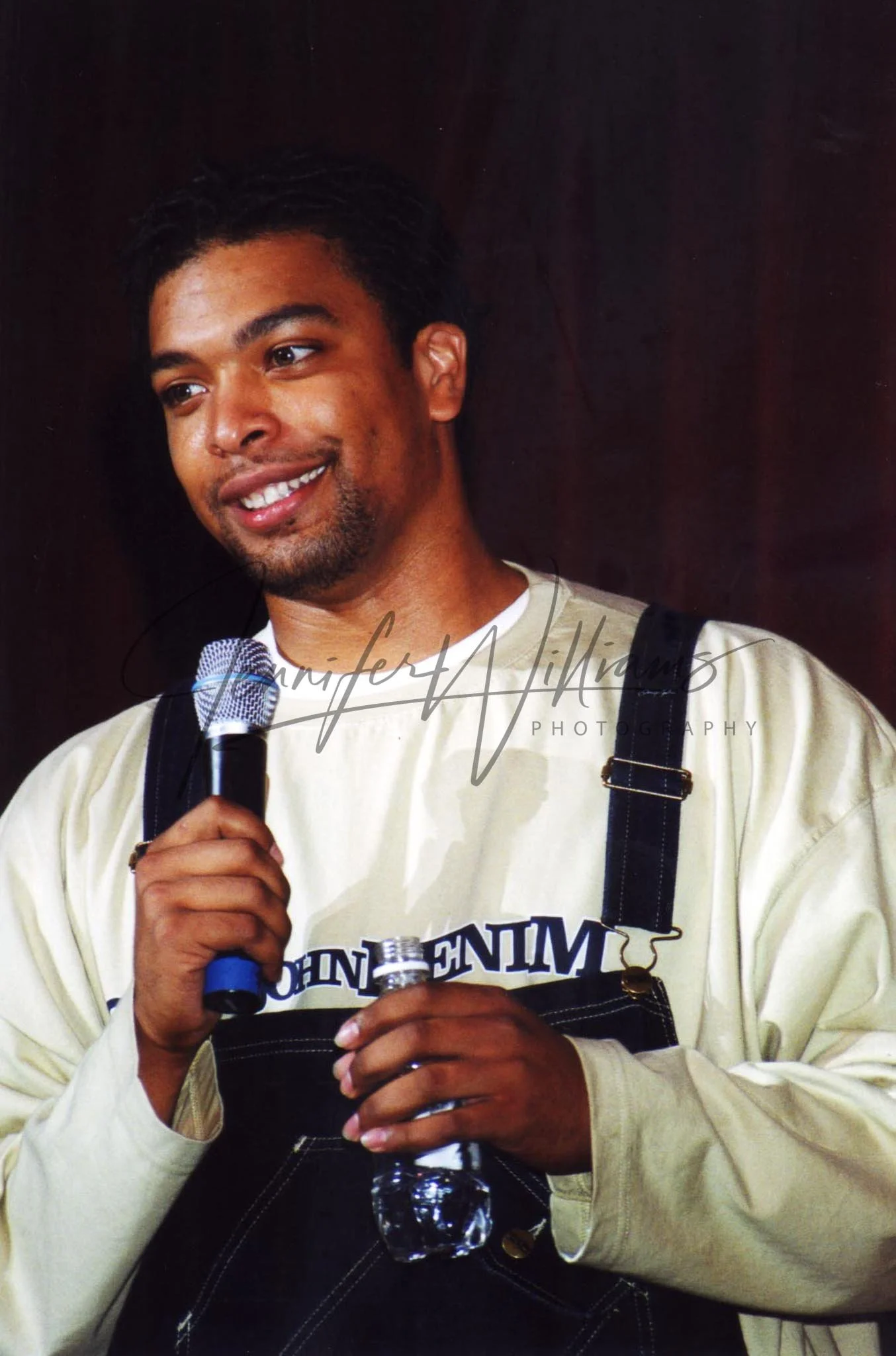 A man holding a microphone and a water bottle, smiling, wearing a white shirt and black overalls, with a dark background.